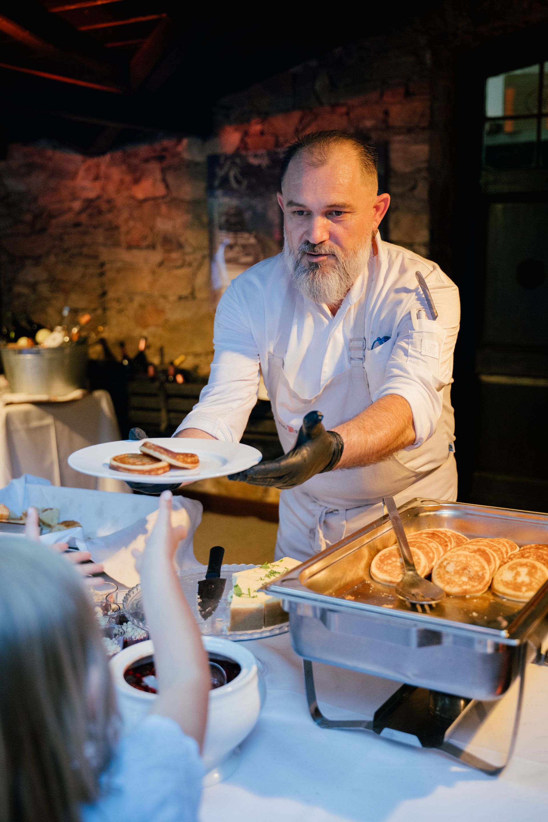 Ein Koch in weißer Uniform serviert einer Person einen Teller mit Essen an einem Buffet in einem Raum mit Steinwänden.