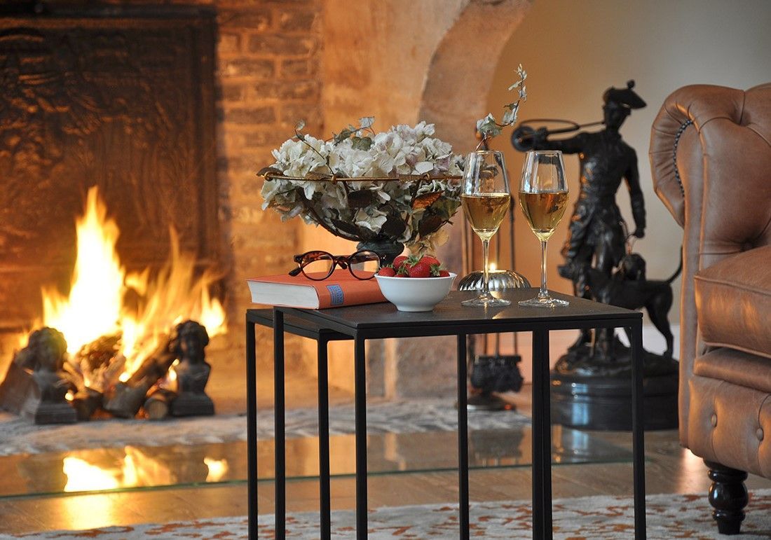 Zwei Gläser Weißwein und eine Schale Erdbeeren stehen auf einem schwarzen Tisch vor einem prasselnden Kaminfeuer.