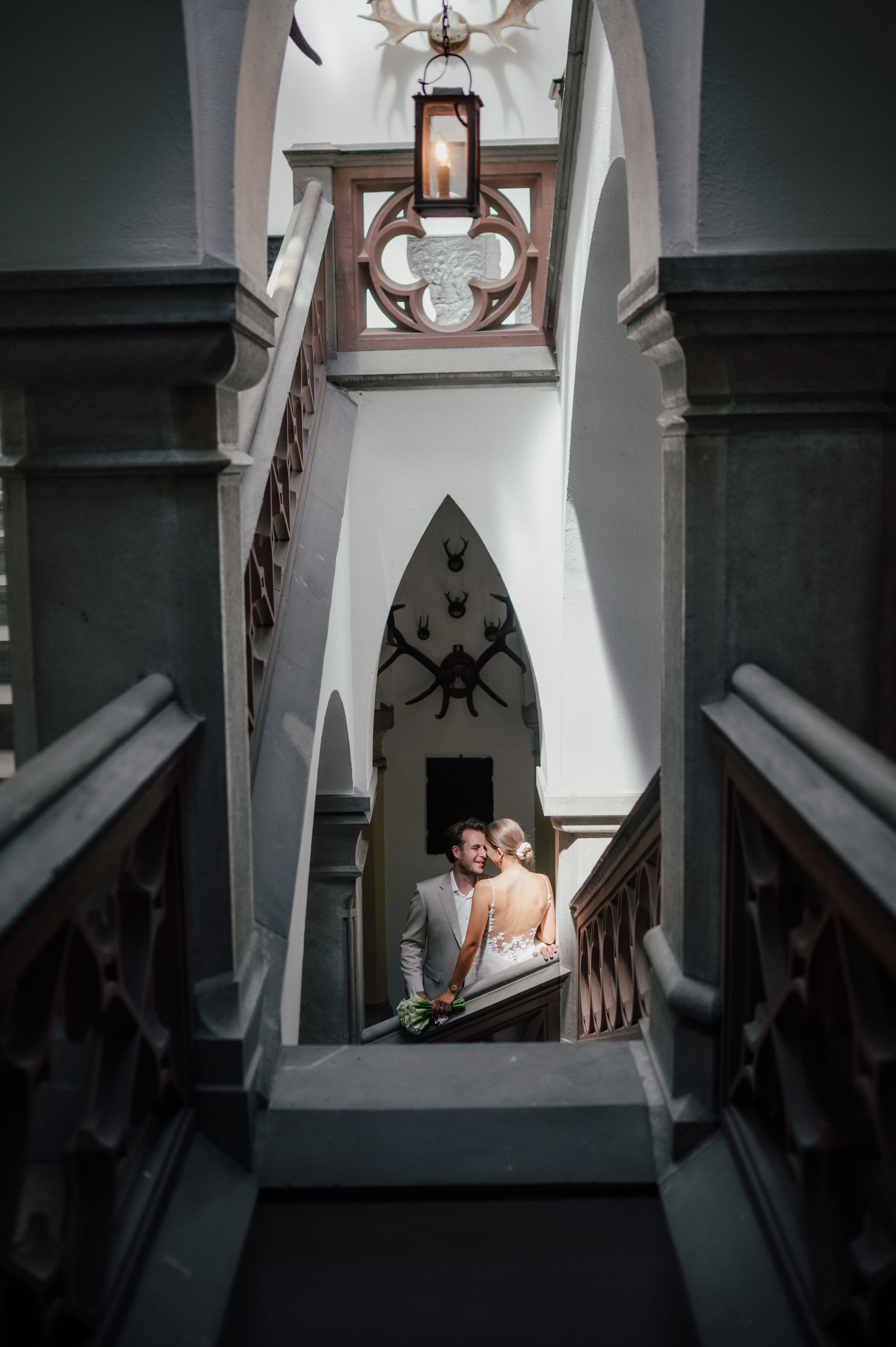 A couple is embracing on an indoor staircase in an ornate, castle-like hallway with Gothic arches and hanging lanterns.