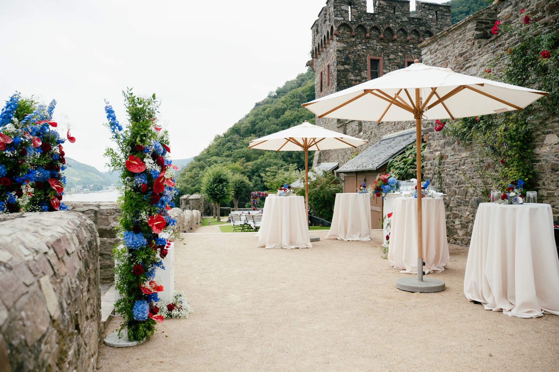 Eine Freiluft-Eventterrasse mit weißen Tischen und Sonnenschirmen vor einer steinernen Burgmauer, die mit roten und blauen Blumen geschmückt ist.