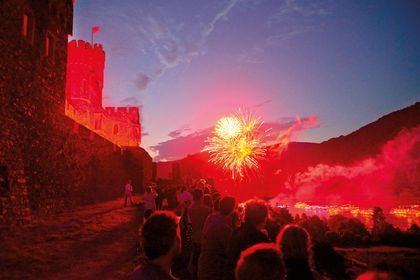 Eine Menschenmenge beobachtet ein Feuerwerk vor dem Abendhimmel in der Nähe einer Burgmauer, die in hellrotem Licht erstrahlt.