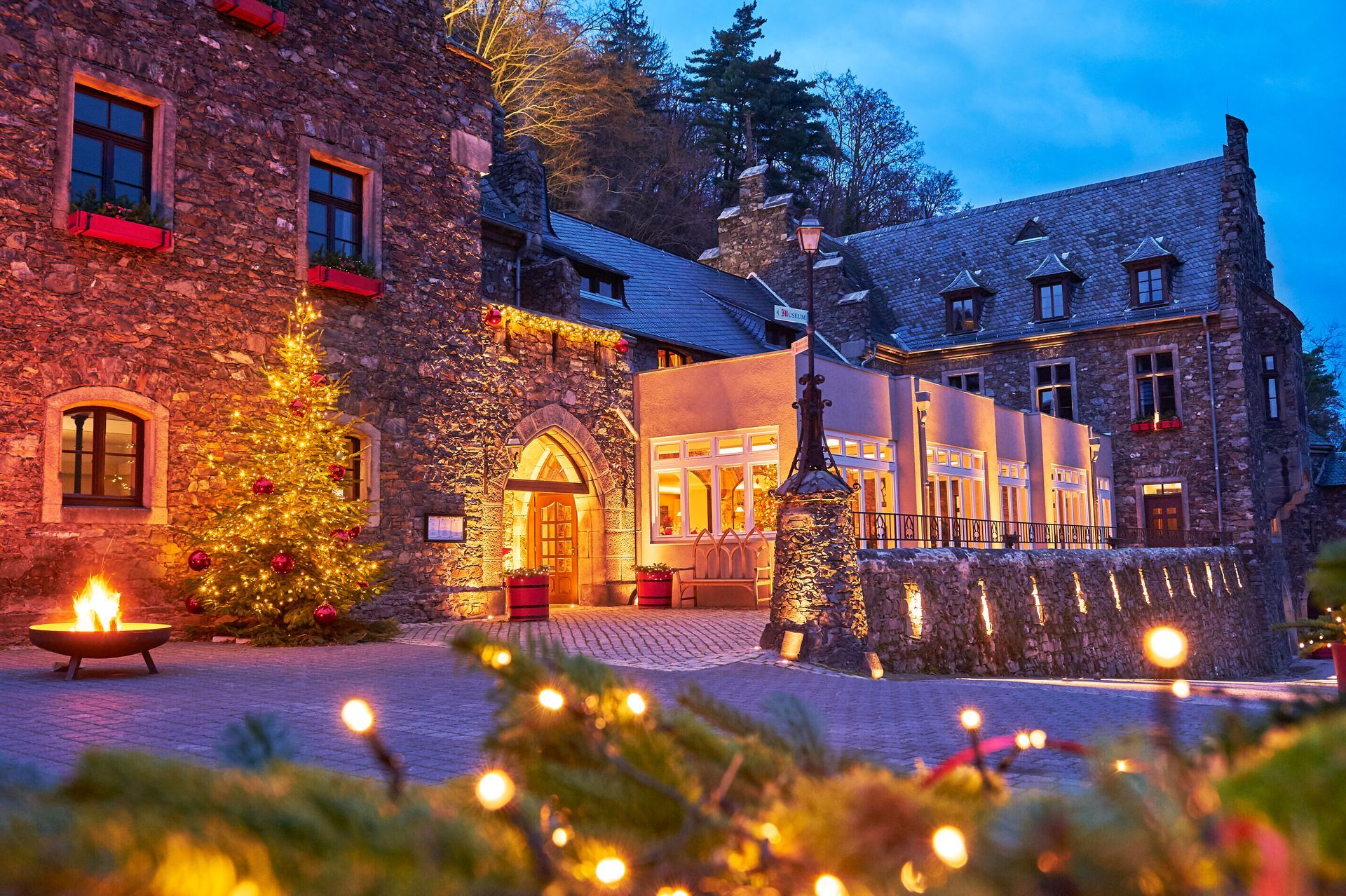 Eine steinerne Burg in der Abenddämmerung, weihnachtlich geschmückt mit einem beleuchteten Baum, warmen Außenlichtern und einer Feuerstelle im Innenhof.
