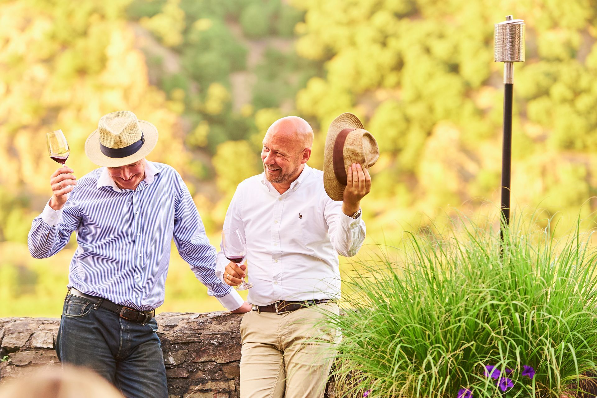 Zwei Männer mit Hüten und Hemden mit Knöpfen halten Weingläser in der Hand und stehen im Freien in der Nähe einer Steinmauer.
