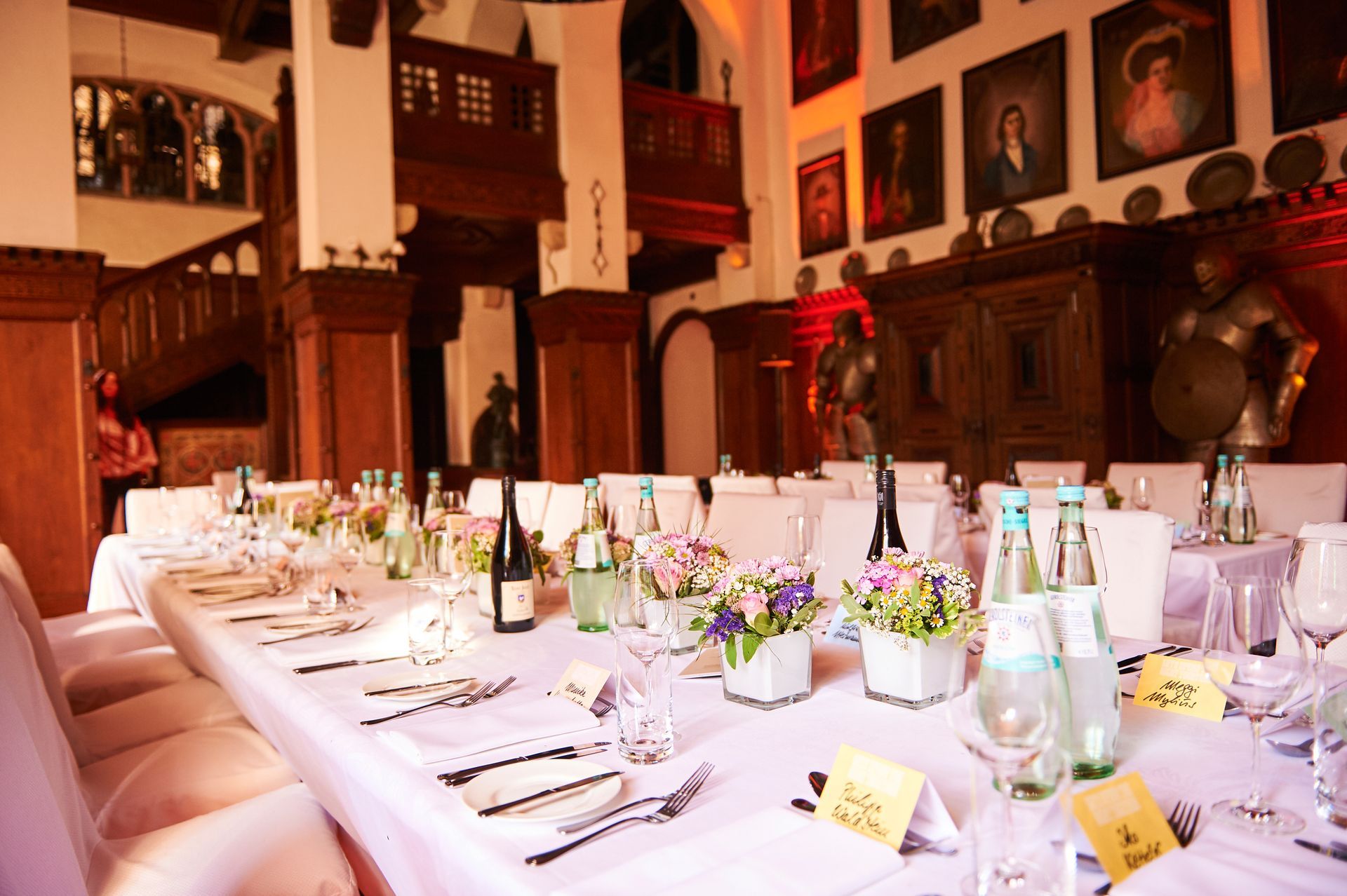 A long banquet table, set for a formal dinner, in a historic room adorned with portraits, dark wood, and decorative suits of armor.