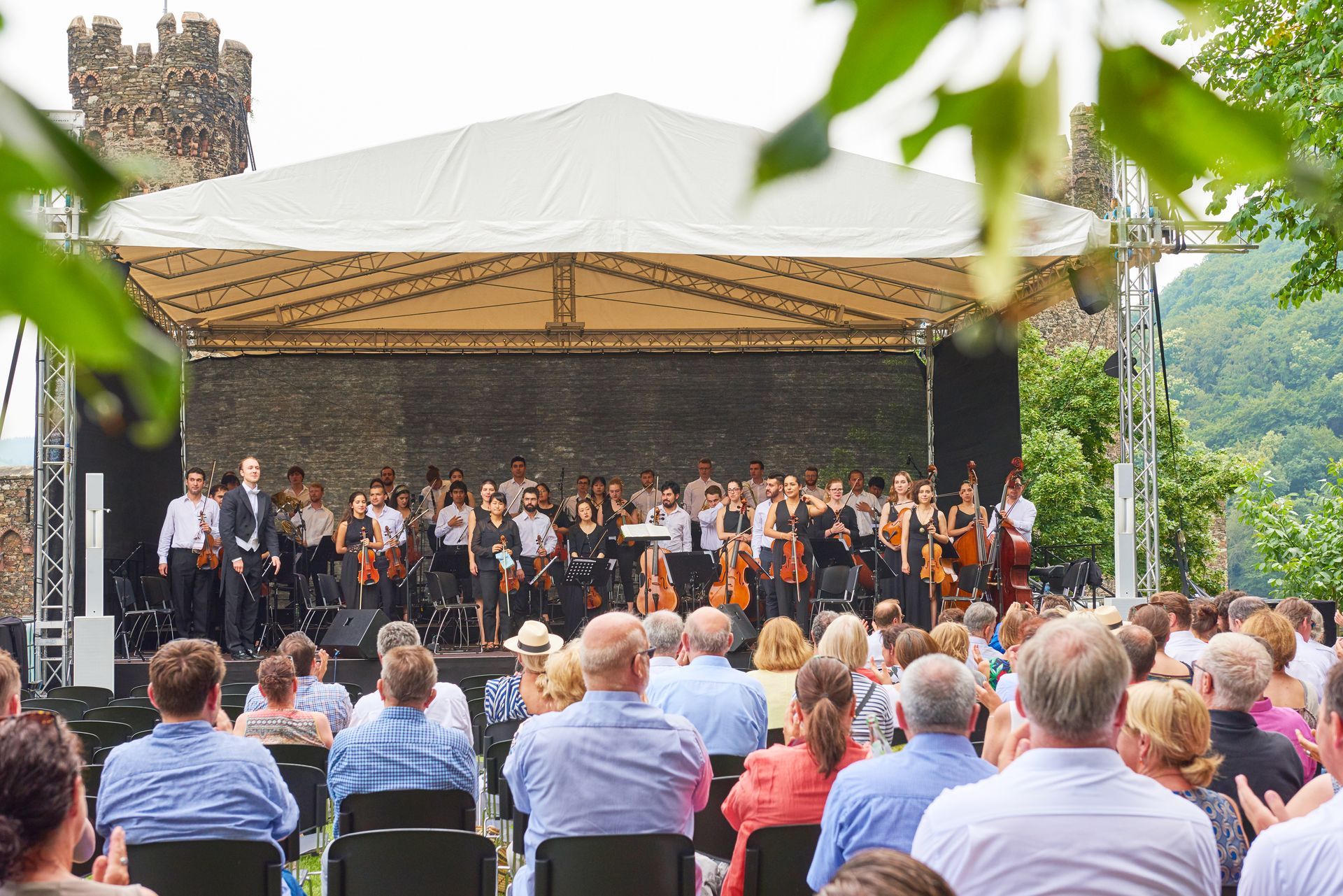 Outdoors, spectators sit and watch an orchestra playing on a covered stage in front of the ruins of a stone castle.