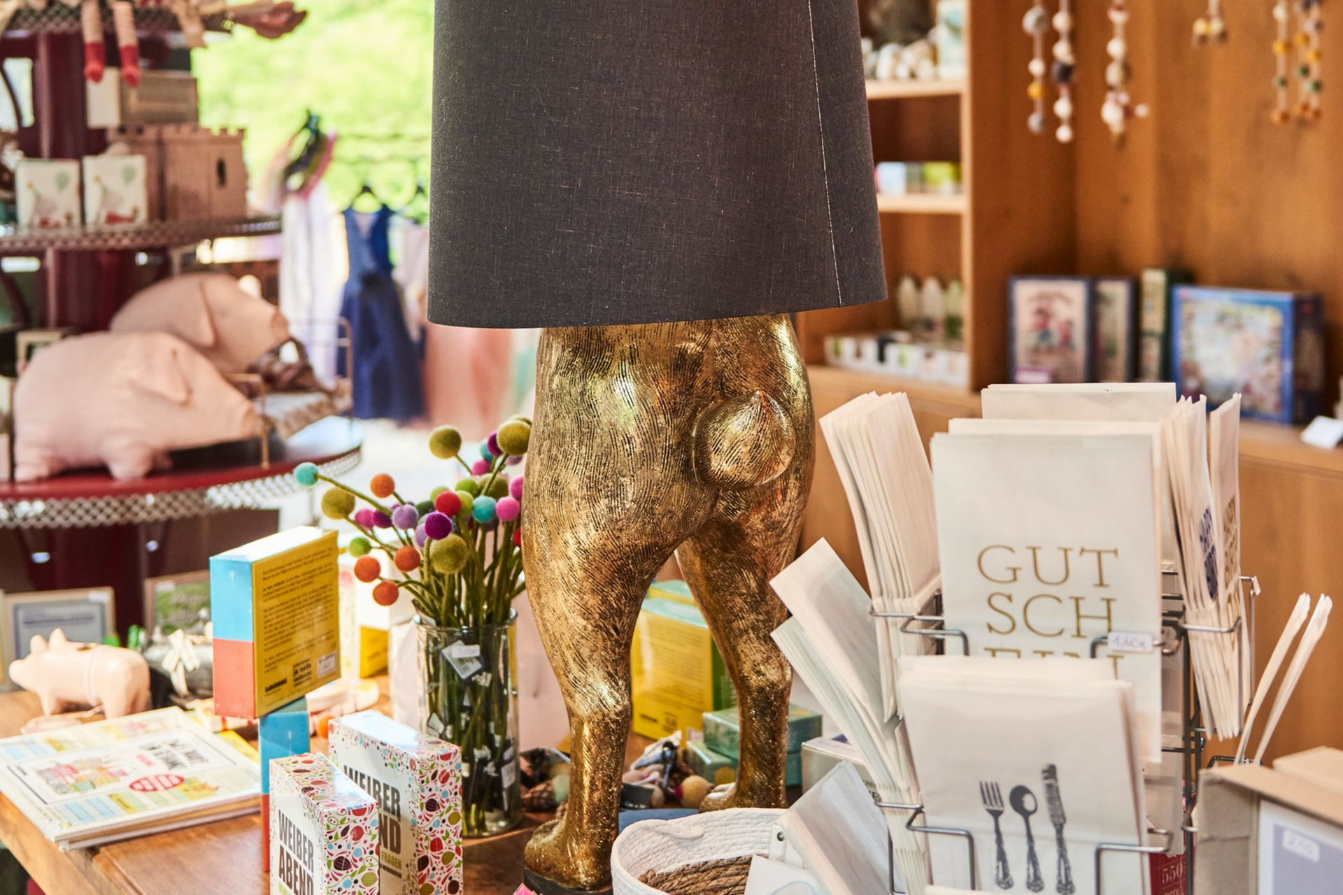 In einem Geschäft ist ein Tisch ausgestellt, auf dem eine Lampe mit einem goldenen, tierförmigen Fuß, farbenfrohe Blumendekorationen und Geschenkkarten zu sehen sind.