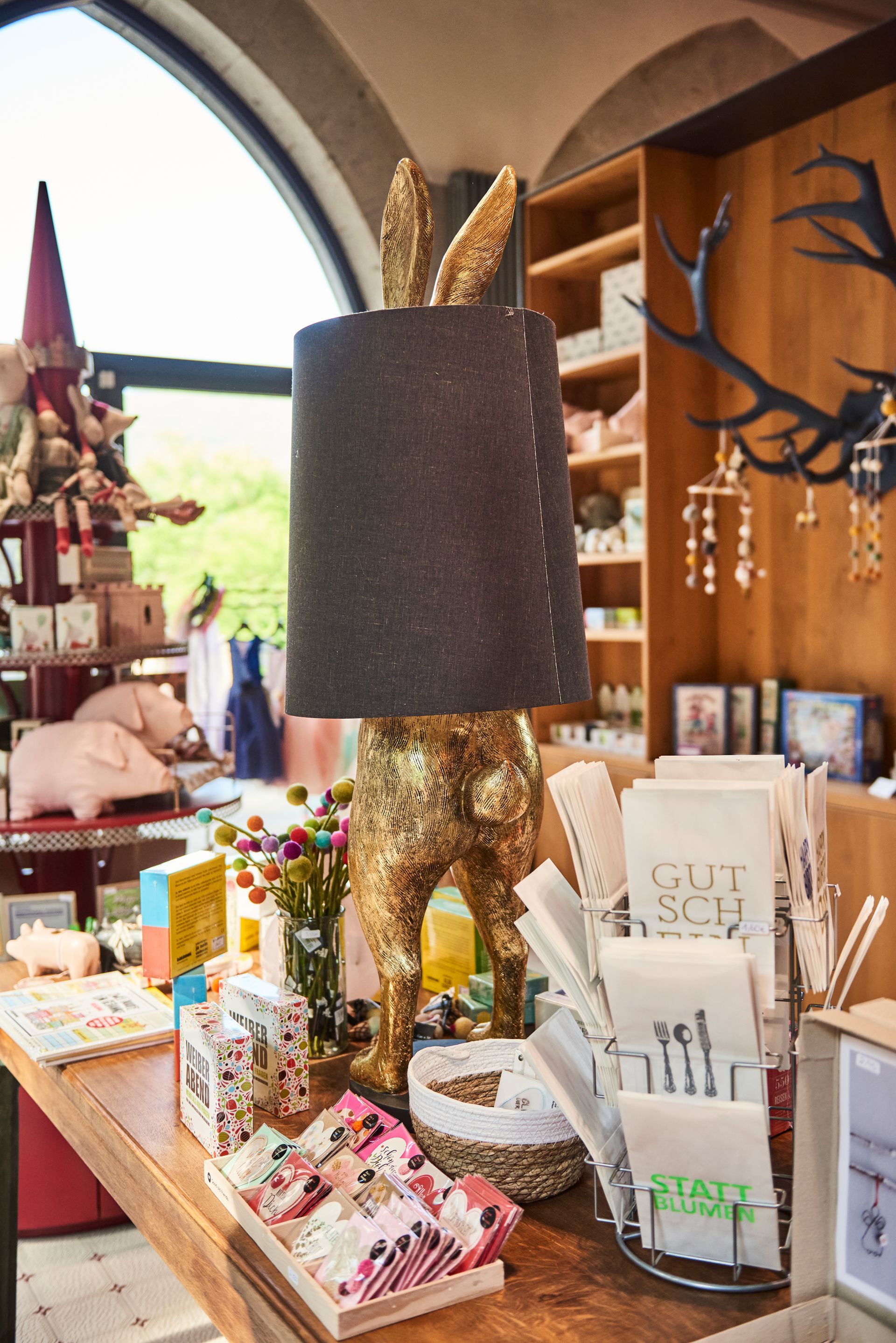 On a display table in a store, a golden rabbit-shaped lamp with a dark shade sits next to gift items and small pig figurines.