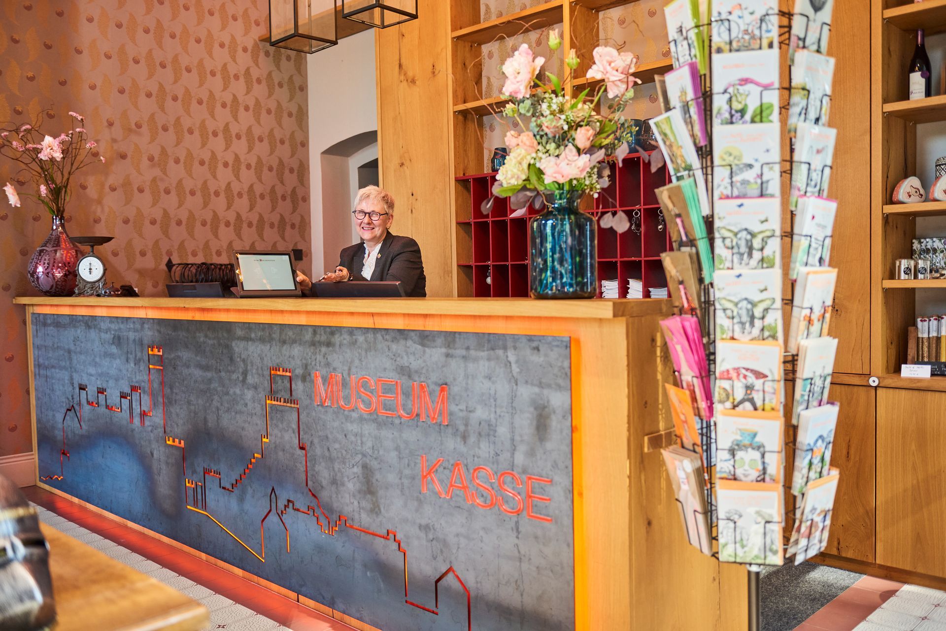 A museum ticket counter attendant; in the background, there is a postcard stand, flowers, and shelves.