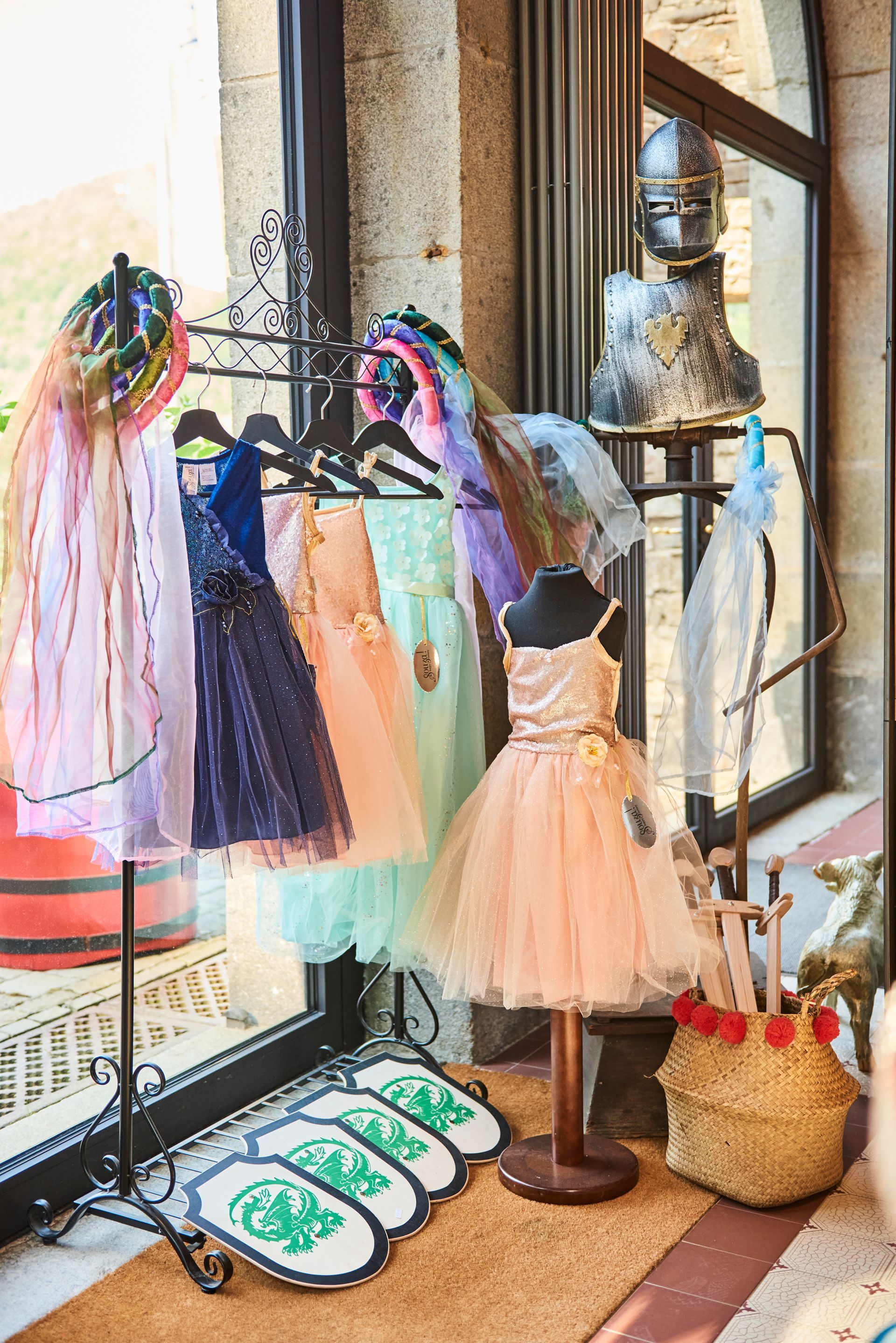 A clothes rack with colorful clothes, silk scarves, and a dress on a mannequin, with shields and a helmet next to it.