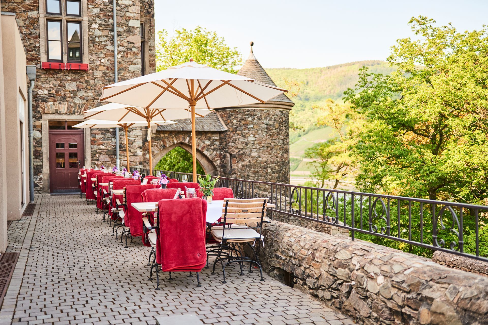 Ein Restaurant mit Außenterrasse und Tischen, die mit roten Tischdecken bedeckt und unter Sonnenschirmen stehen, neben einer historischen Steinmauer.