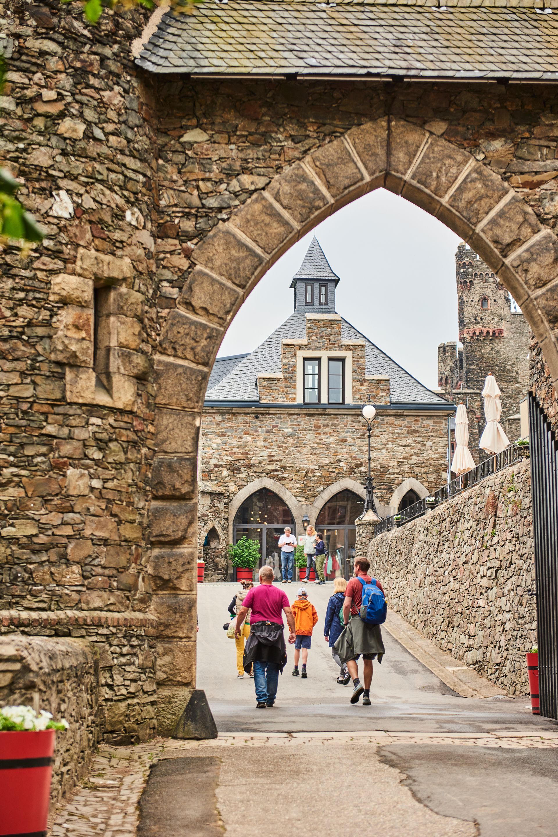 Die Menschen schreiten durch einen steinernen Torbogen, der zu einem historischen Gebäude mit grauem Dach und Steinmauern führt.