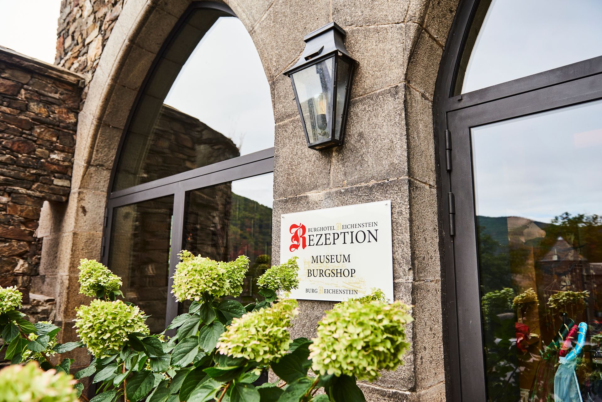 Ein Schild mit der Aufschrift „Rezeption“ neben einem gläsernen Bogeneingang eines Steingebäudes mit grünen blühenden Sträuchern im Eingangsbereich.