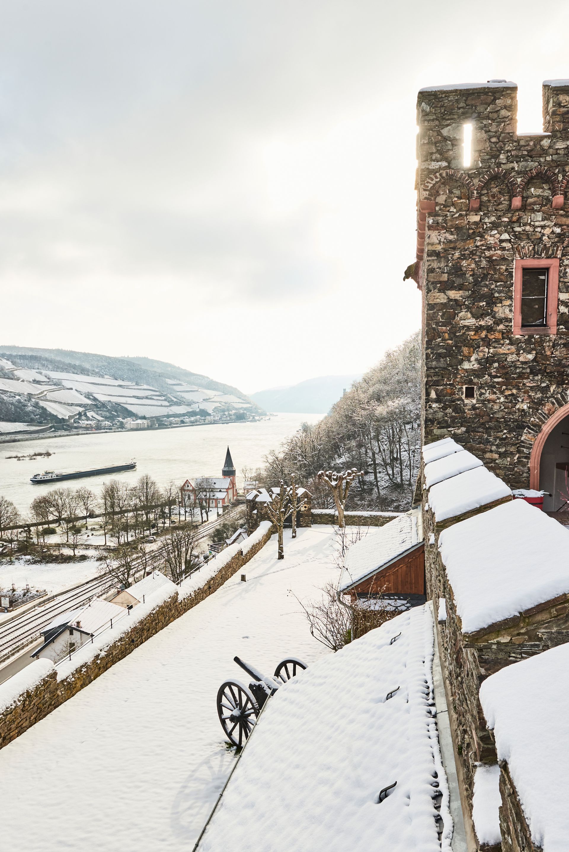 Eine schneebedeckte Burgmauer aus Stein mit einer alten Kanone überblickt ein zugefrorenes Flusstal und ferne, schneebedeckte Hügel.