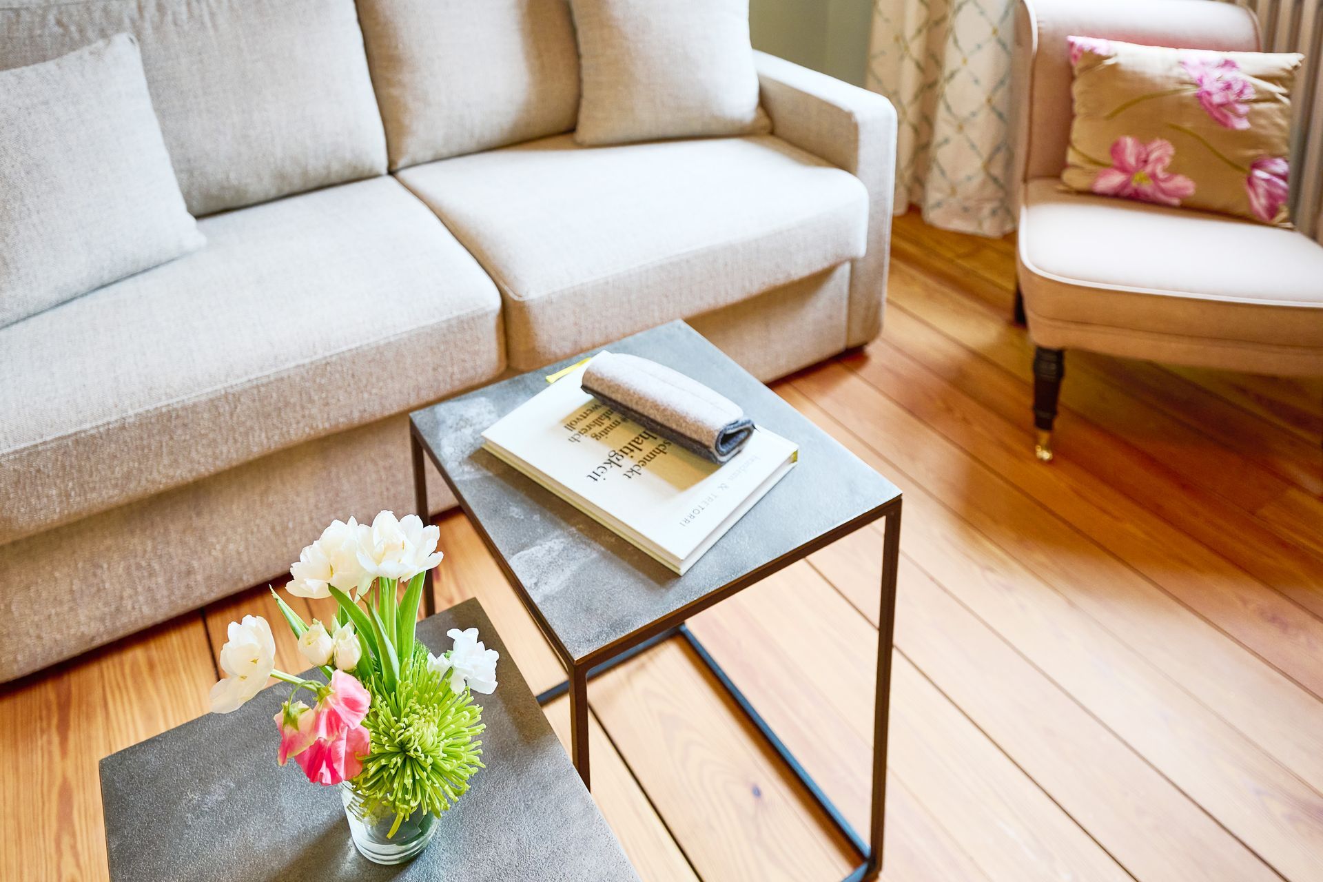 A beige sofa, an armchair with a floral pattern, and two dark coffee tables, one of which has a vase of flowers and a book on it.