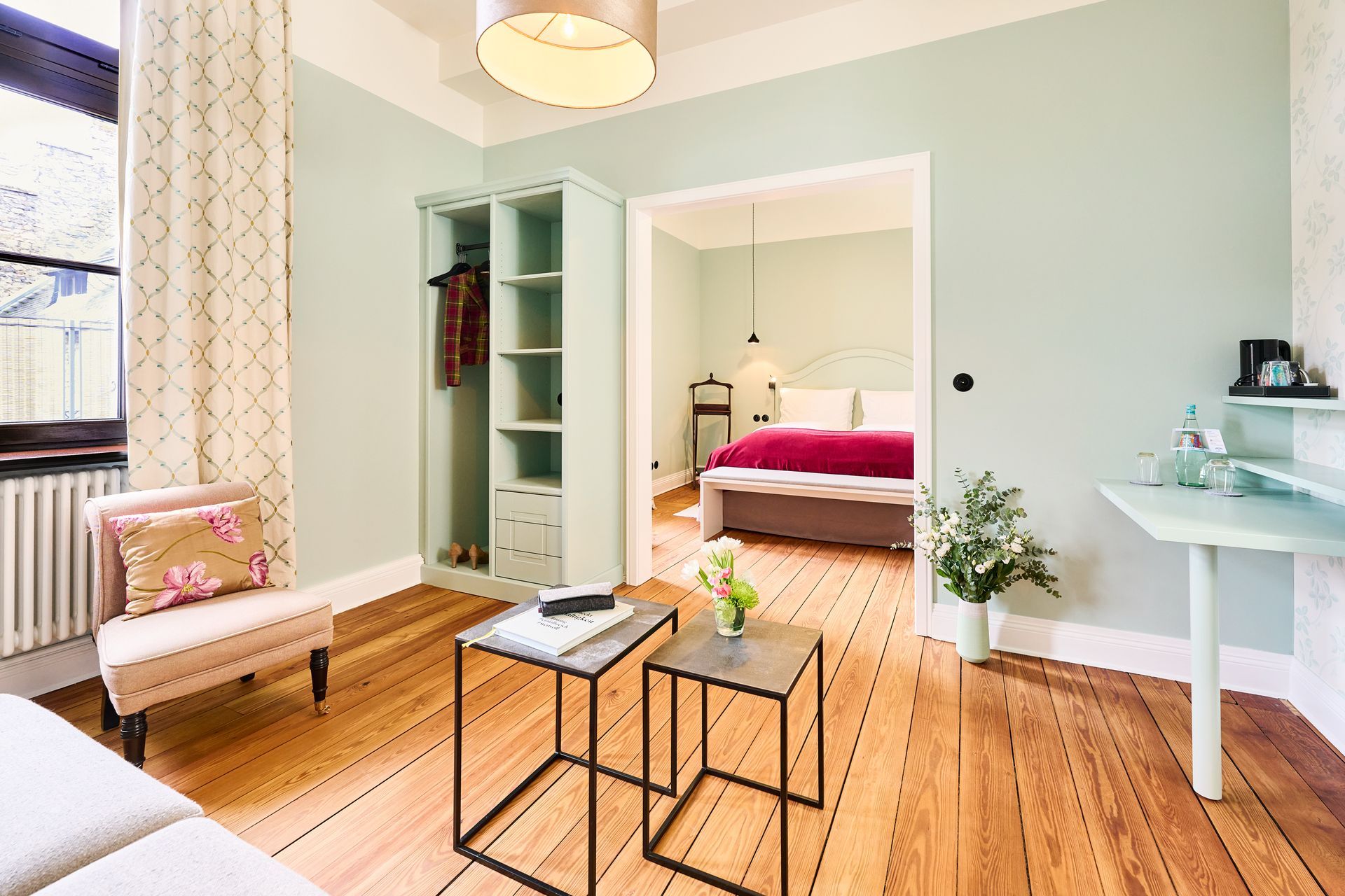 A brightly lit hotel suite with light green walls, hardwood floors, a small sitting area, and a view into the bedroom.