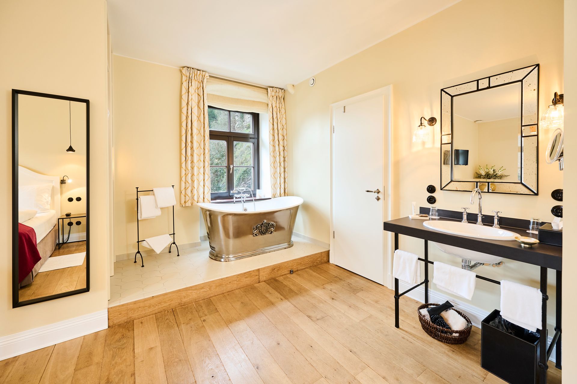 A bright, airy hotel bathroom with hardwood floors, a freestanding metal bathtub near the window, and a modern vanity mirror.
