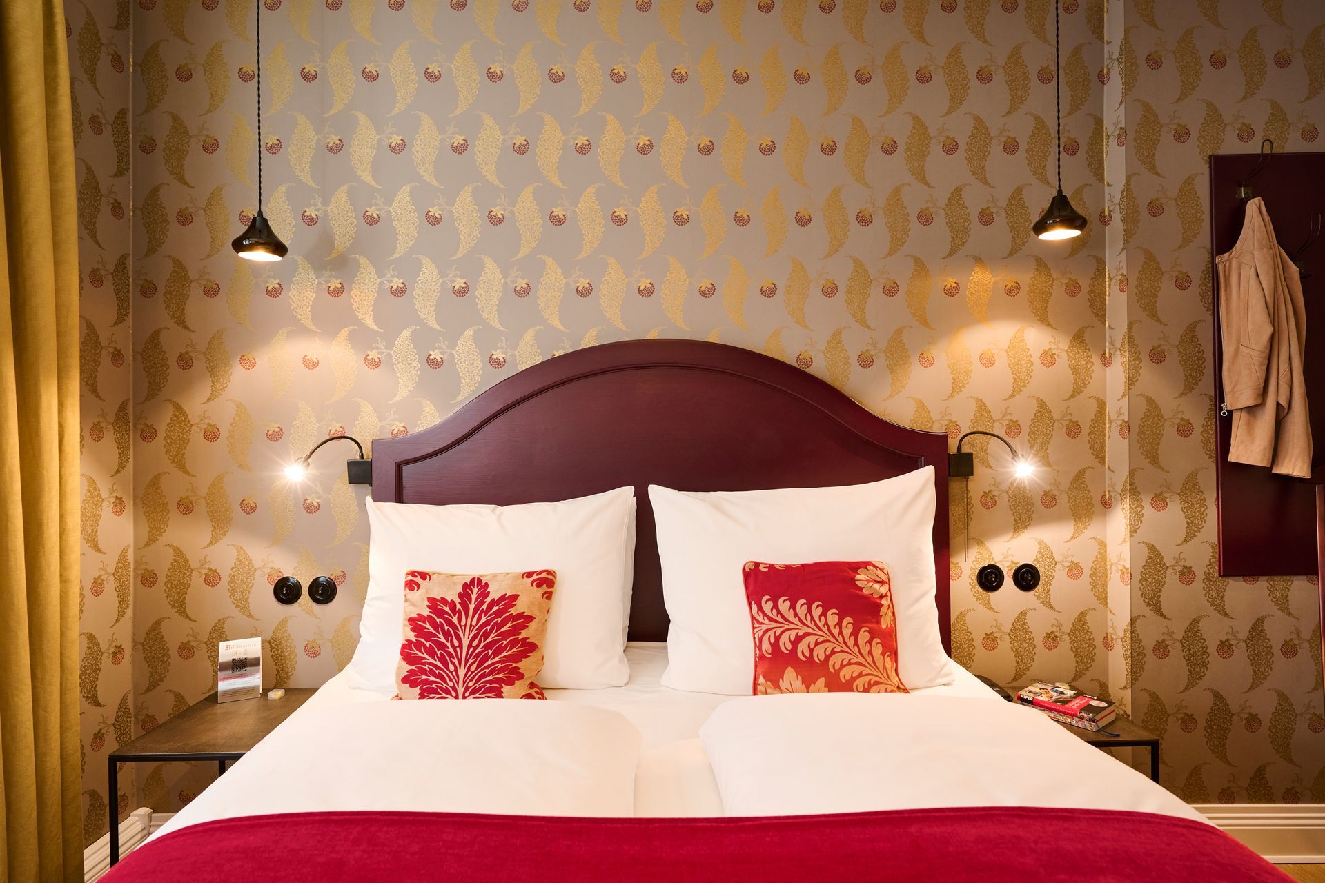 A bedroom with a dark purple headboard, patterned wallpaper, pendant lights, and a red bedspread on a white bed.