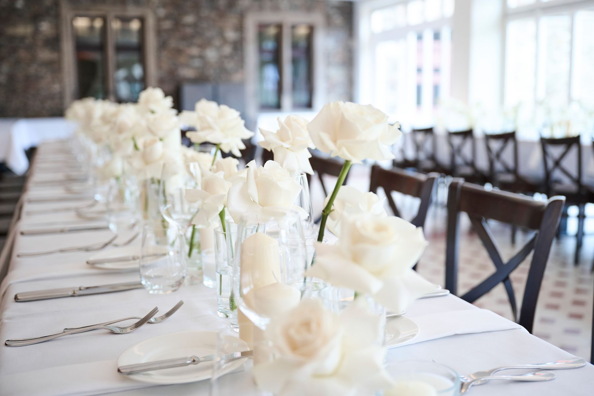 Eine lange Tafel, gedeckt für einen festlichen Anlass, mit weißen Rosen in Glasvasen und Gedecken auf einer weißen Tischdecke.