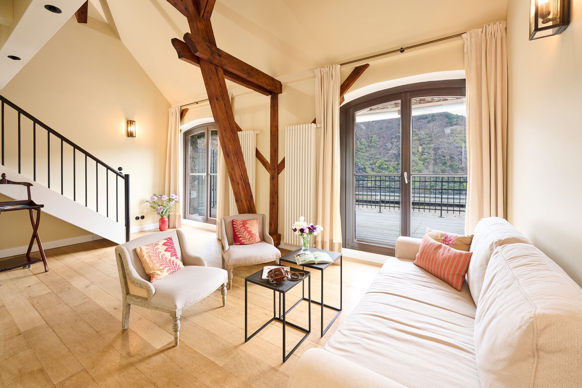 Helles Hotelzimmer mit weißem Sofa, Sesseln, Holzbalken und großem Bogenfenster mit Blick auf eine Terrasse.