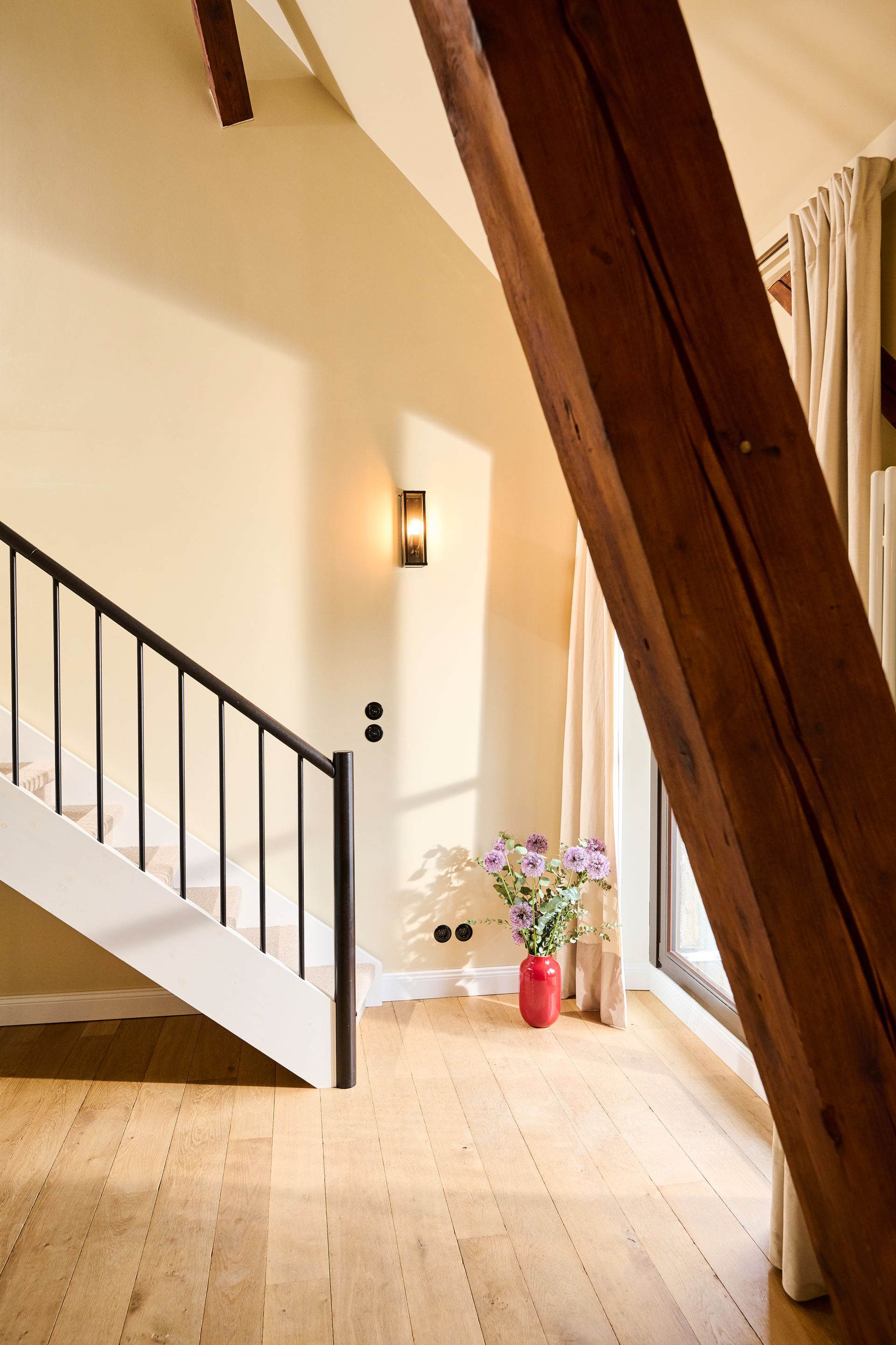 An interior view of a bright room with a wooden floor, a modern staircase, a wall sconce, and a red vase of flowers.