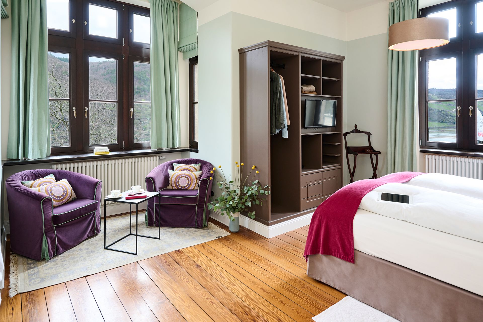A hotel room with two purple armchairs, a small table, a wooden wardrobe, and a bed with a pink bedspread.