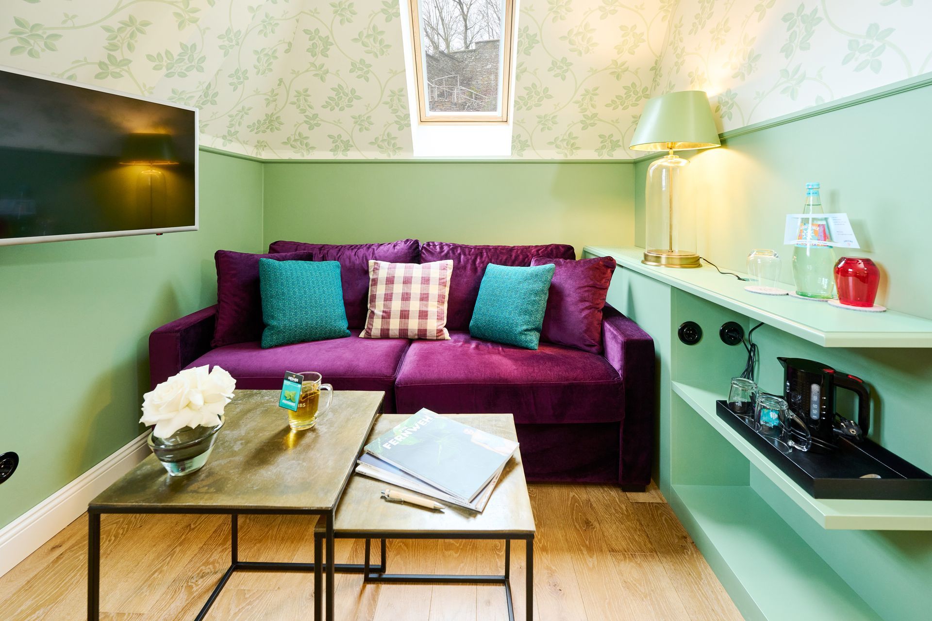 Ein lila Sofa mit türkisfarbenen und gemusterten Kissen steht vor einer hellgrünen Wand in einem Zimmer mit Fernseher und Kaffeestation.