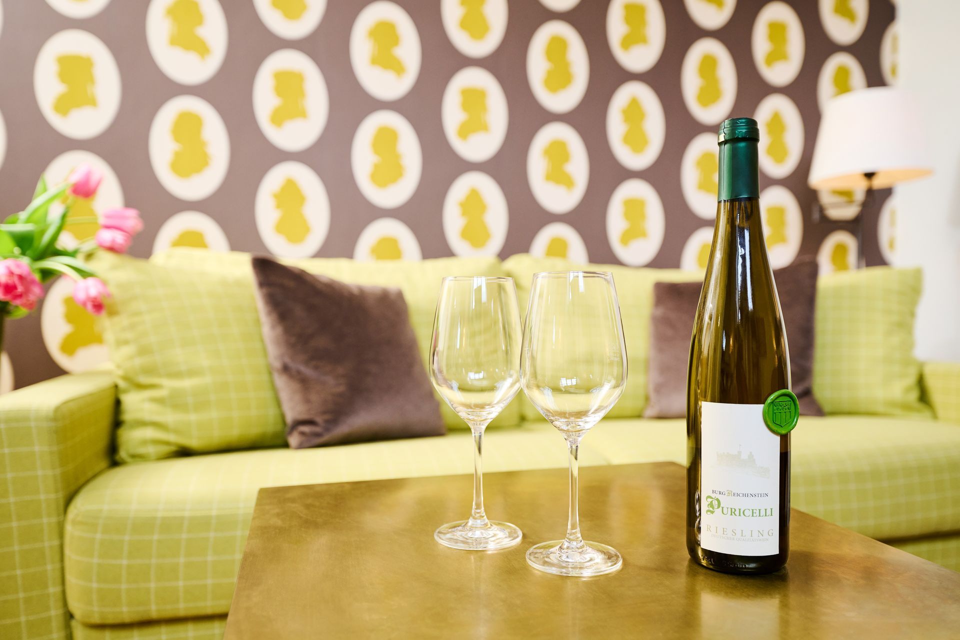 Zwei Weingläser und eine Flasche Weißwein auf einem Couchtisch vor einem grünen Sofa mit einer Wand mit Silhouettenmuster.