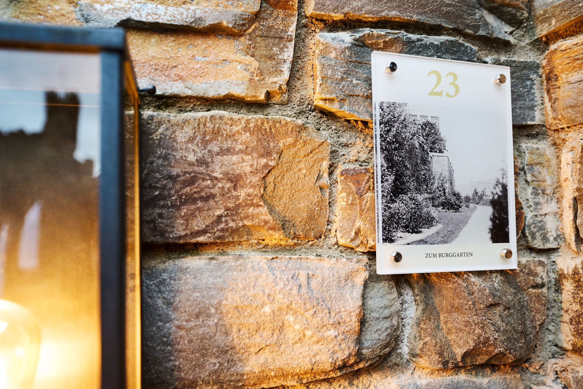 Ein Schwarzweißdruck mit der Aufschrift „23“ ist an einer strukturierten, hellbraunen Steinmauer neben einer beleuchteten Außenlampe angebracht.