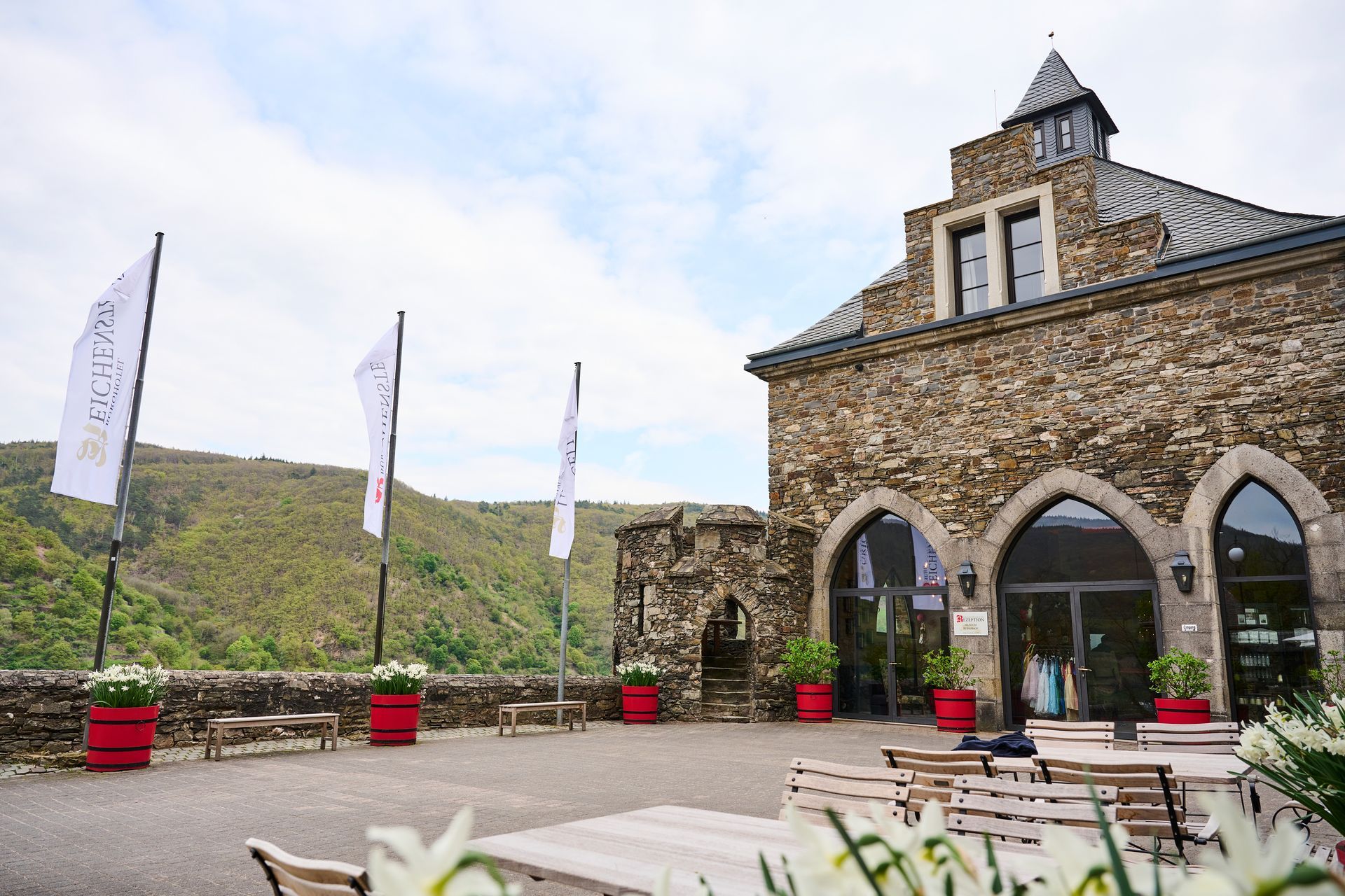 Ein Steingebäude mit Bogentüren und roten Pflanzkästen auf einer Kiesterrasse mit Blick auf einen grünen Hang mit weißen Steinplatten.