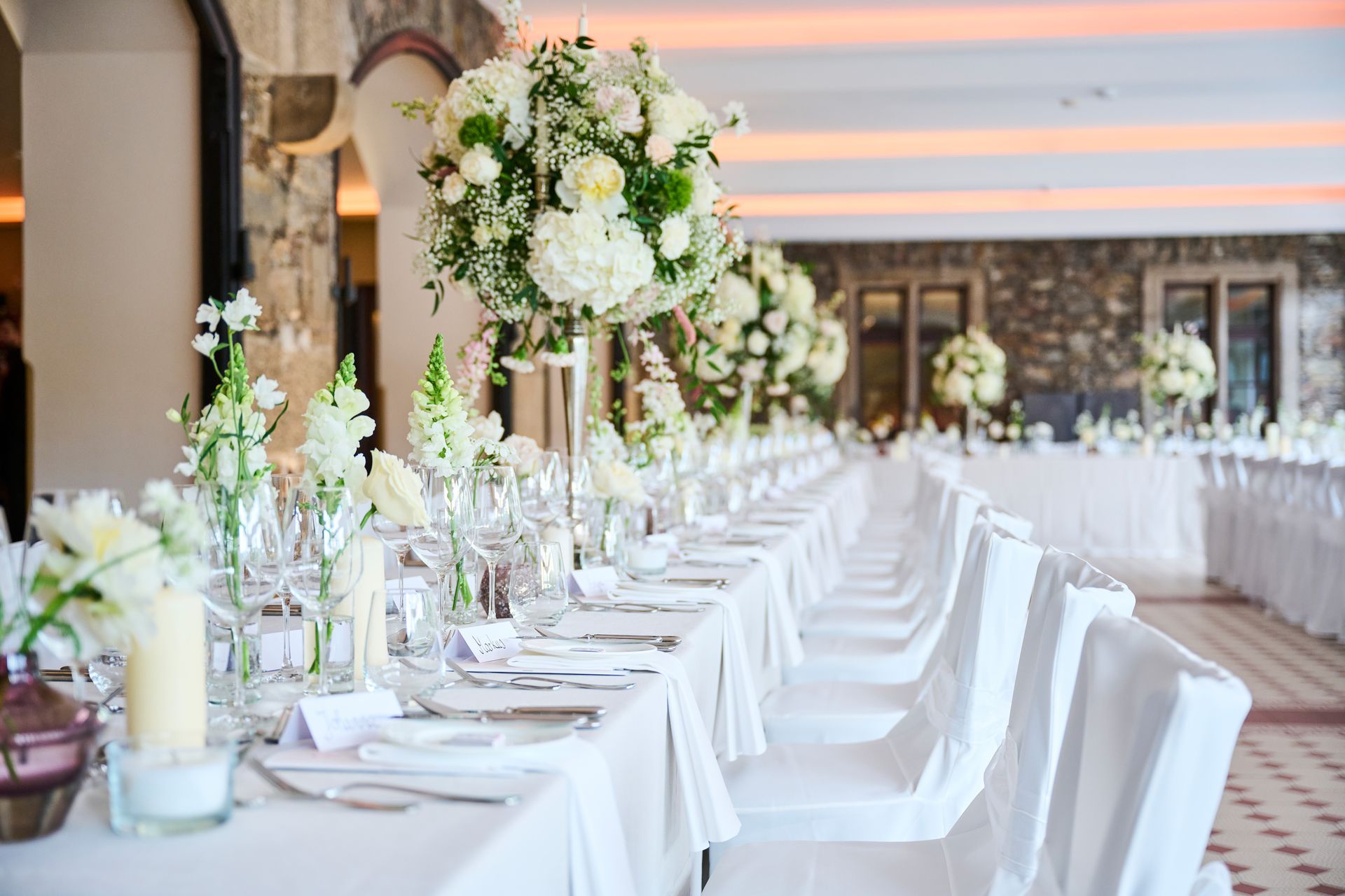 A long wedding table with white tablecloths, chairs, and elegant floral arrangements in a hall surrounded by stone walls.