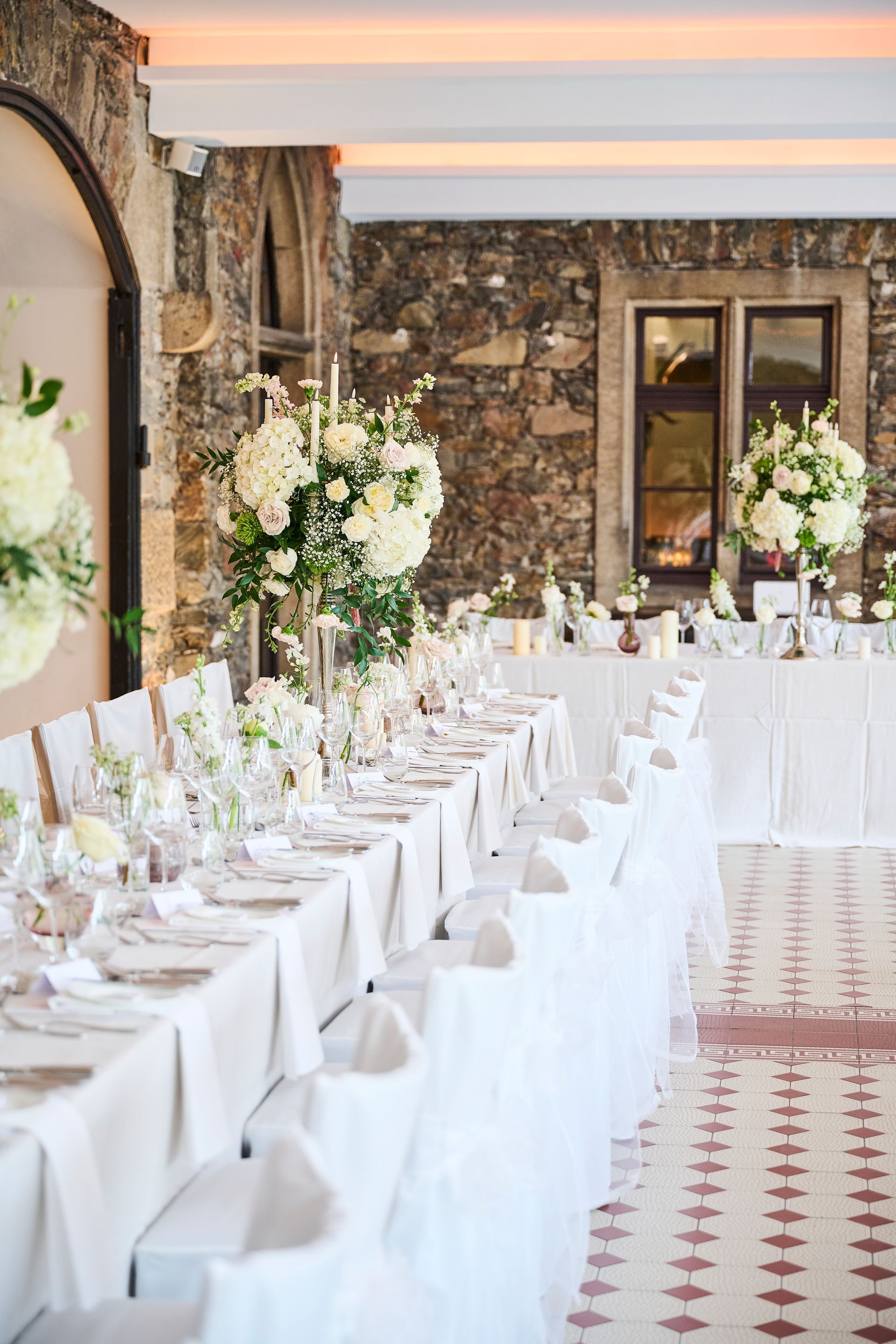 Long banquet tables with white tablecloths, floral arrangements, and upholstered chairs in a room with stone walls.
