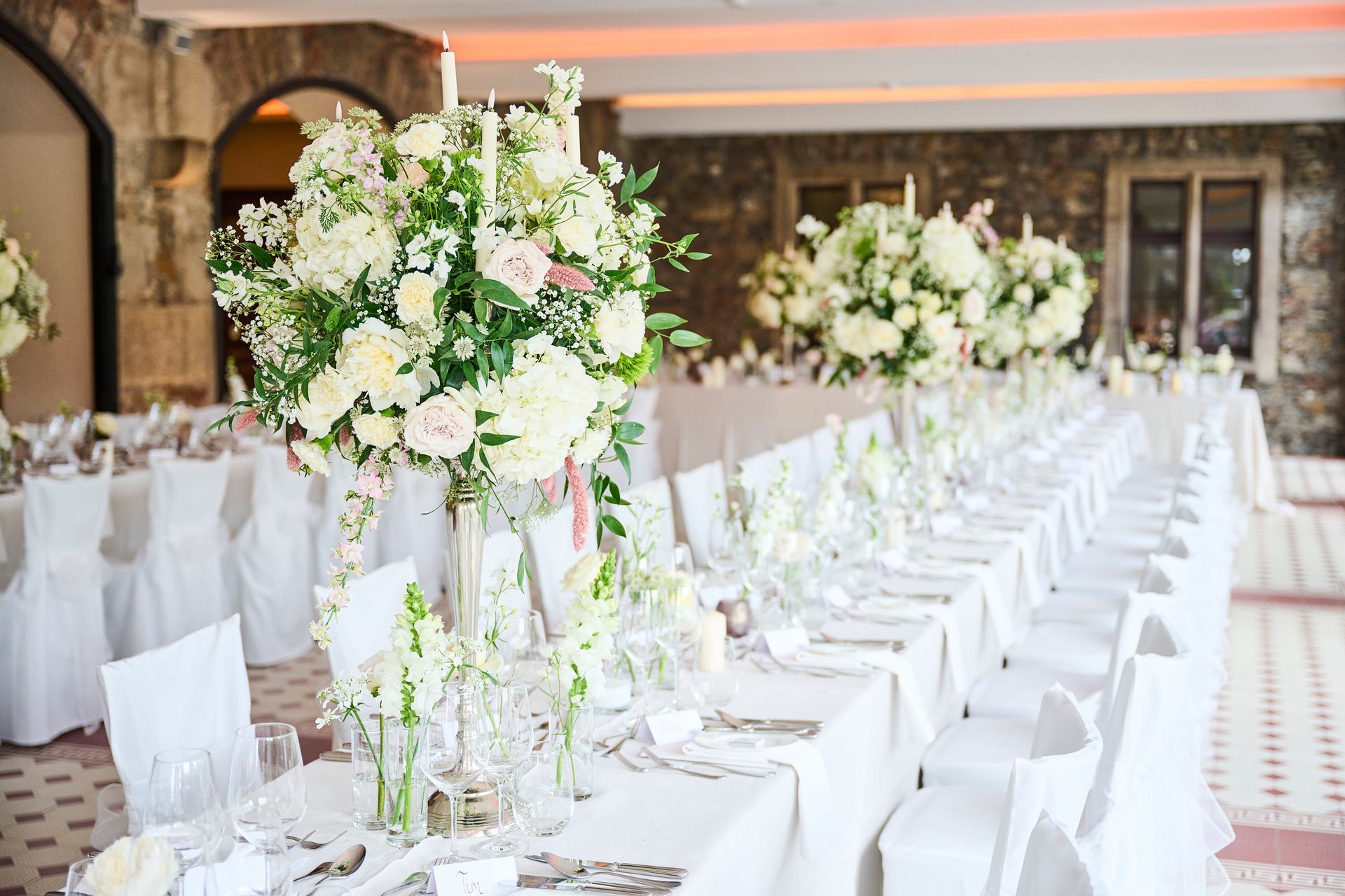 A long banquet table, set for a formal event, with tall white floral arrangements and white chair covers.