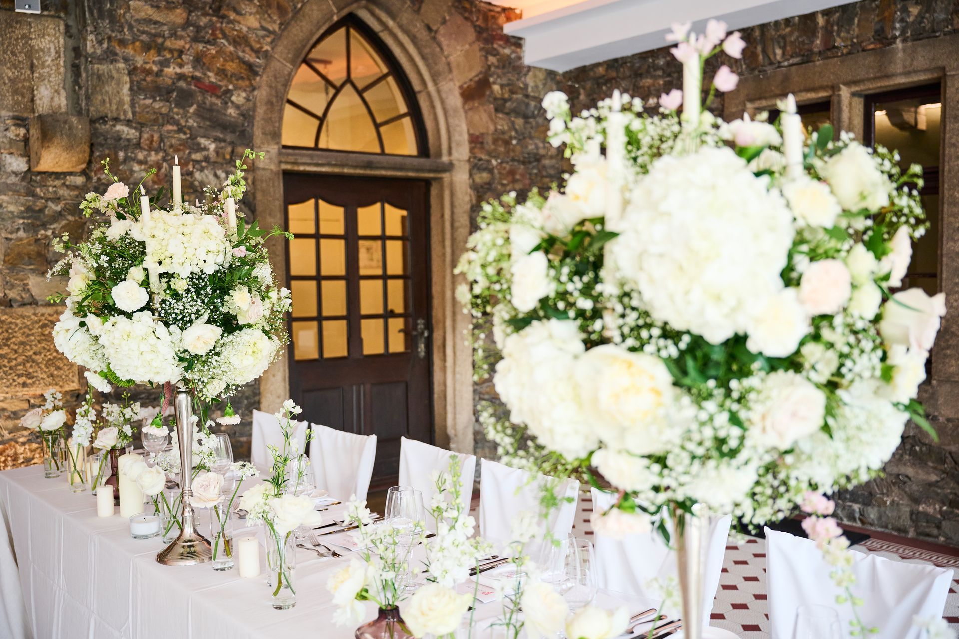 For a formal event, a long table is set with white tablecloths and decorated with large white floral arrangements and candles.