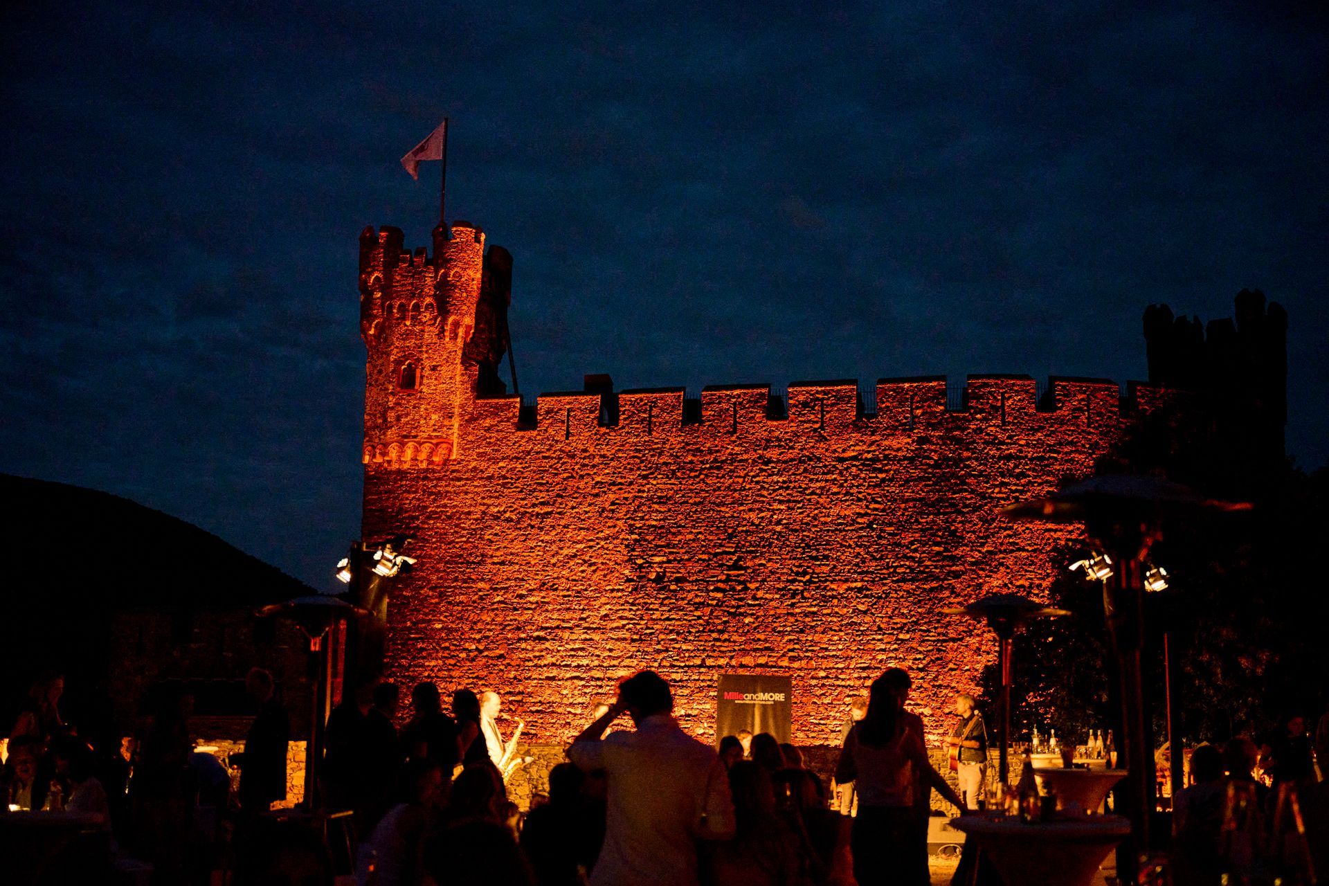 A stone castle, bathed in warm orange light at night, with the silhouettes of people gathered in the foreground.