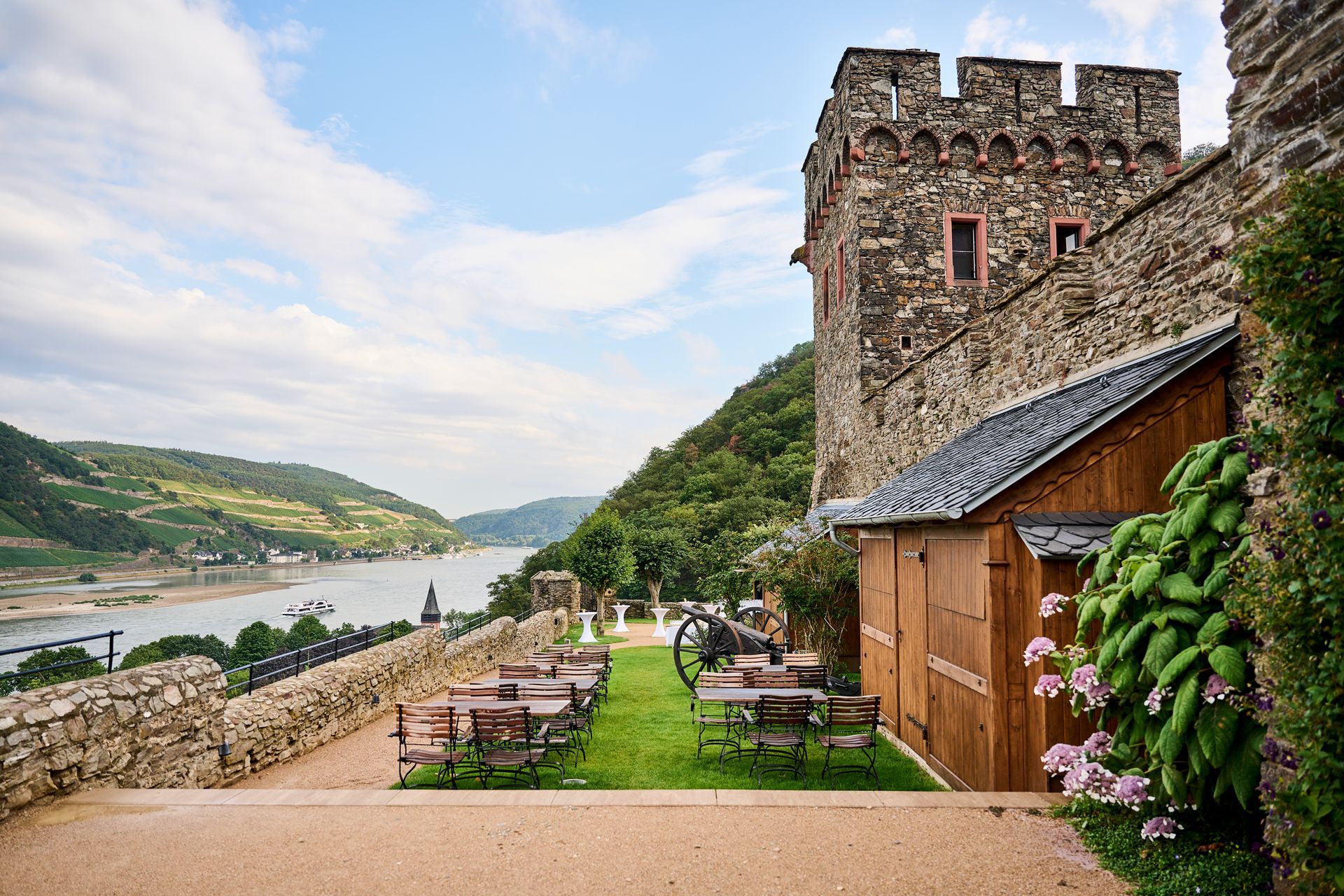 A stone tower and walls overlooking a river valley, with outdoor tables on a grassy terrace.