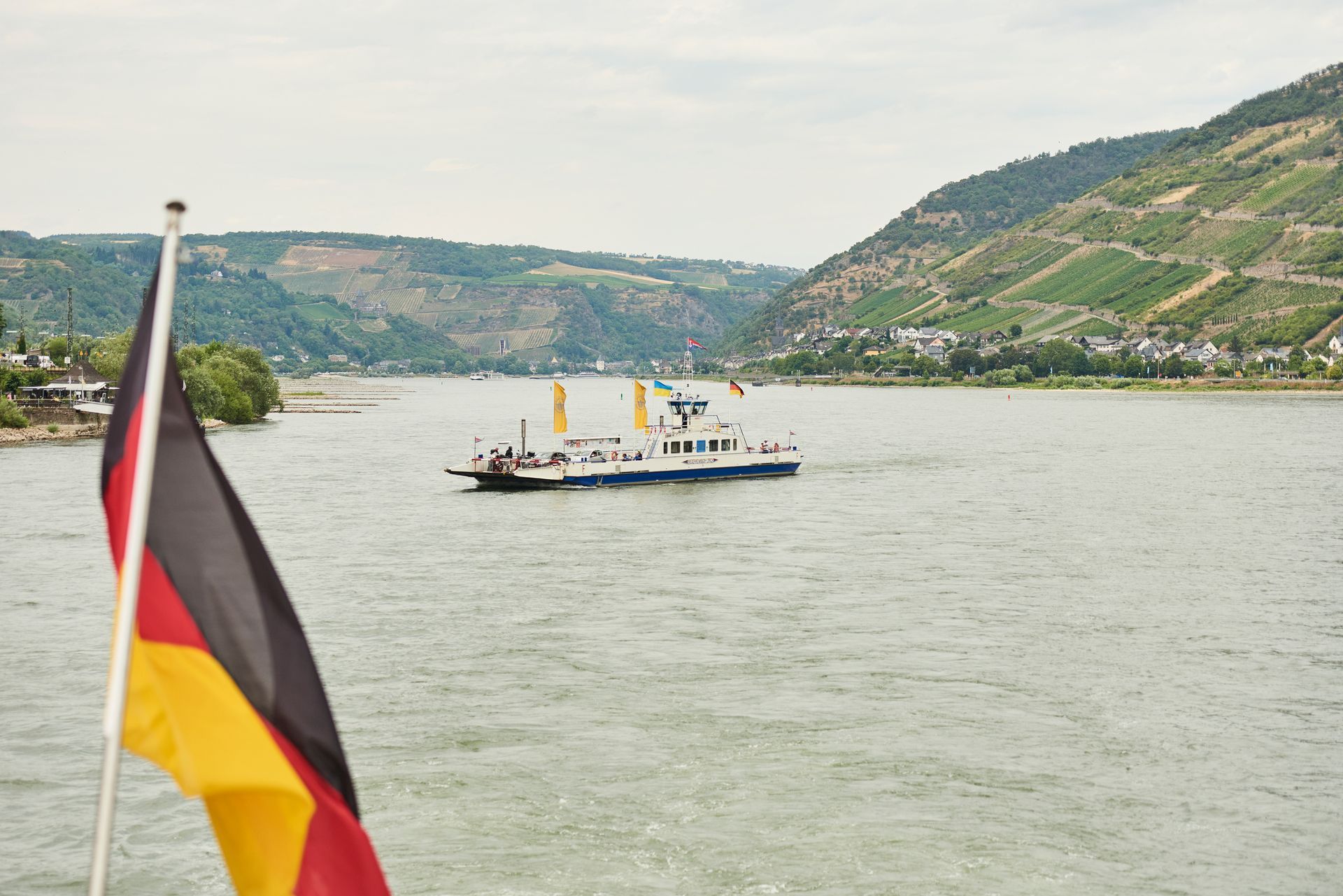 Eine Fähre überquert einen breiten Fluss in Deutschland; im Hintergrund sind Hügel und Weinberge zu sehen, vorbei an einer wehenden deutschen Flagge.