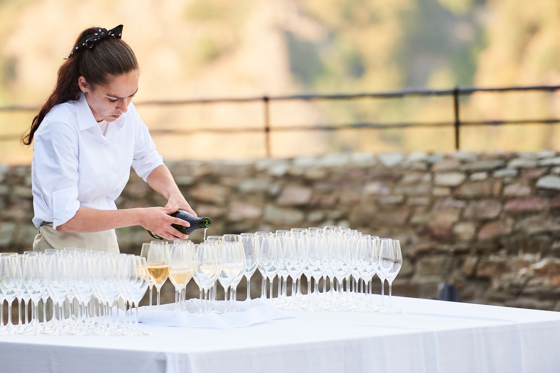 A waiter in a white shirt is pouring champagne into a long row of glasses on a table set up in front of a stone wall in the background.
