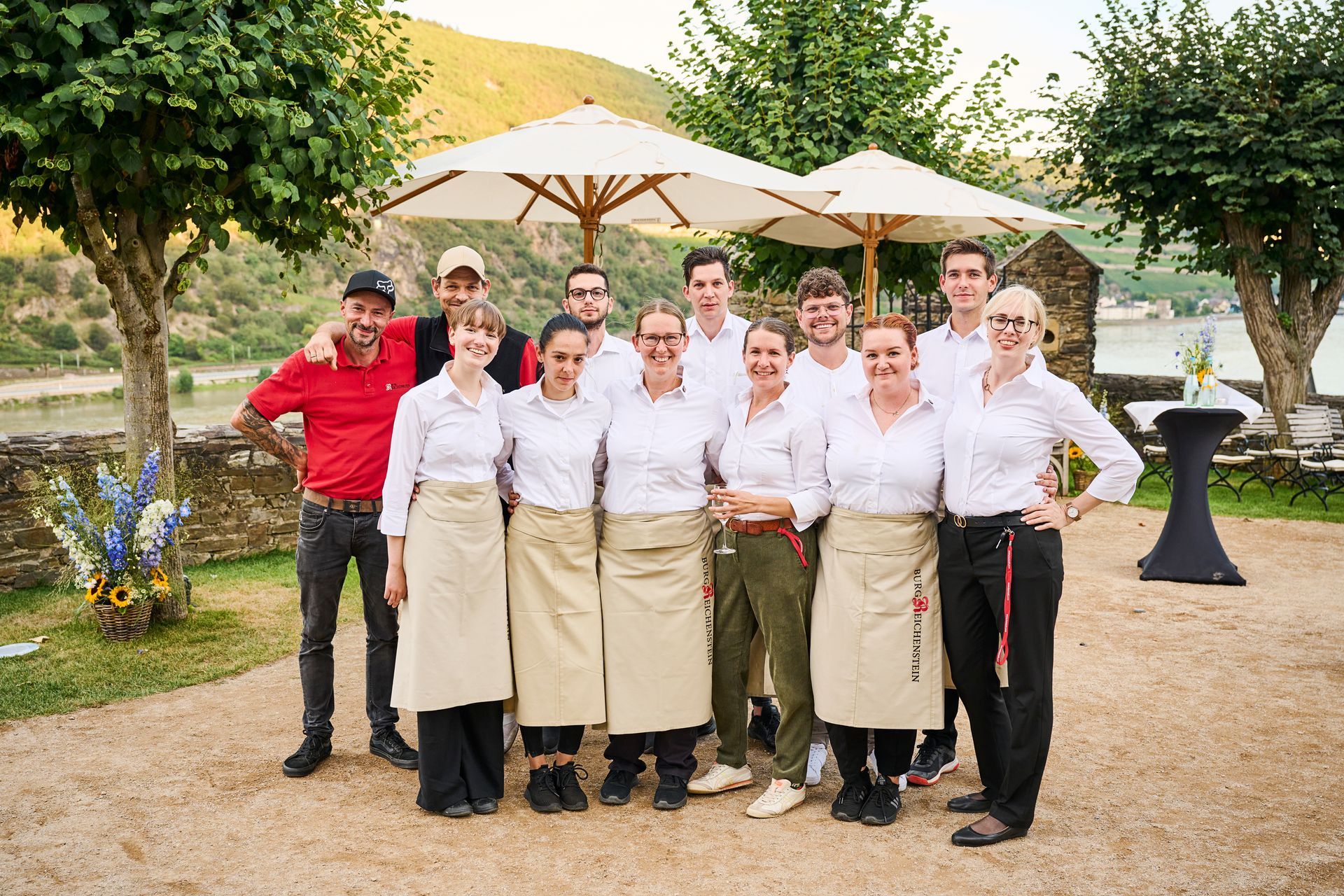 Eine Gruppe Kellner in weißen Hemden und Schürzen steht im Freien unter Sonnenschirmen und lächelt in Richtung eines malerischen Weinbergs.