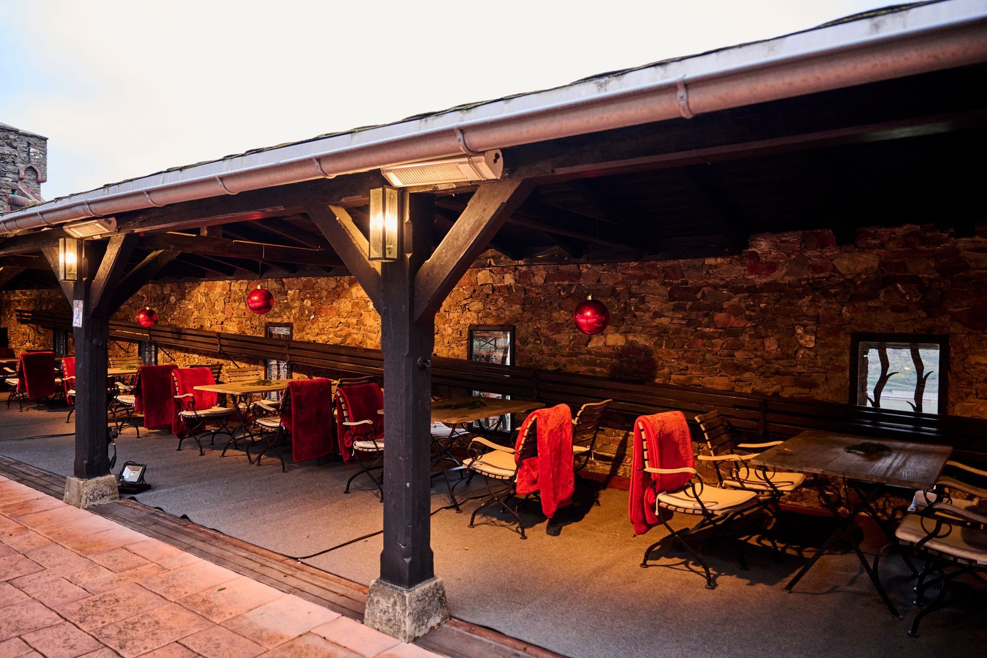 Eine überdachte Terrasse im Freien mit rustikalen Steinmauern, hölzernen Stützbalken und Reihen von Tischen mit Stühlen, die mit rotem Tuch bedeckt sind.