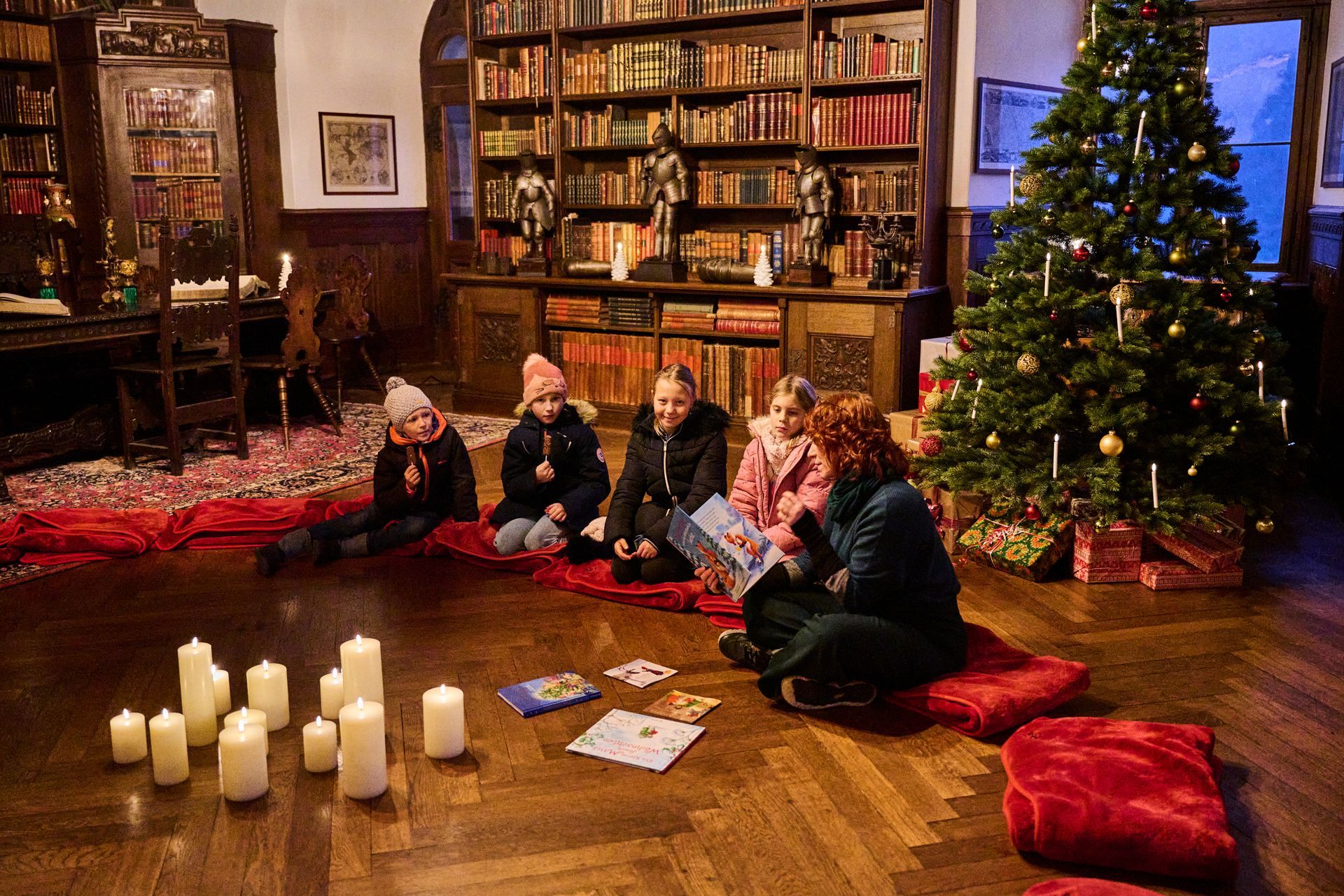 Ein Erwachsener liest vier Kindern vor, die in einer mit Kerzen und einem Weihnachtsbaum geschmückten Bibliothek auf dem Boden sitzen.