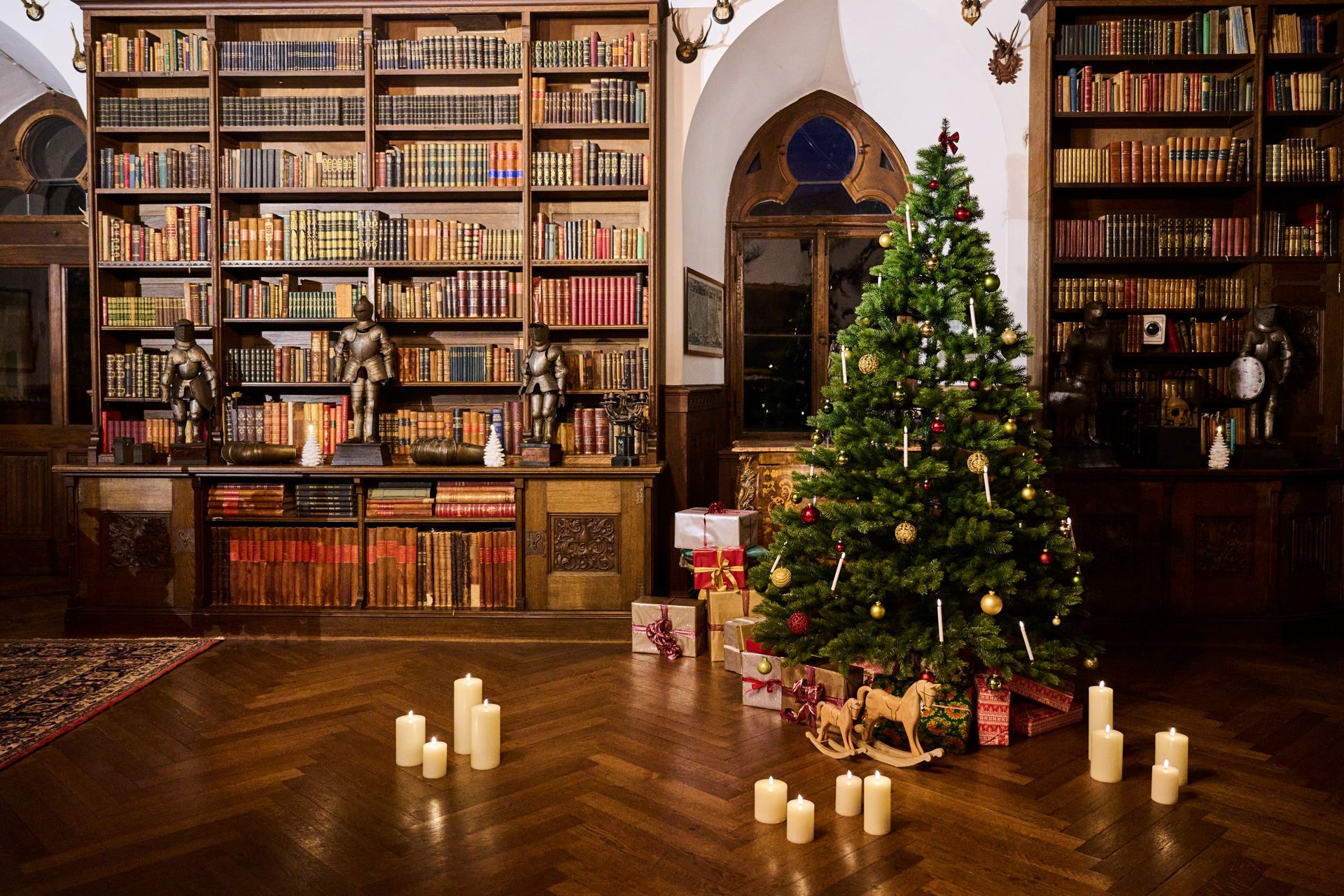 In einer Bibliothek mit raumhohen Bücherregalen steht ein geschmückter Weihnachtsbaum, der von mehreren Stehkerzen beleuchtet wird.