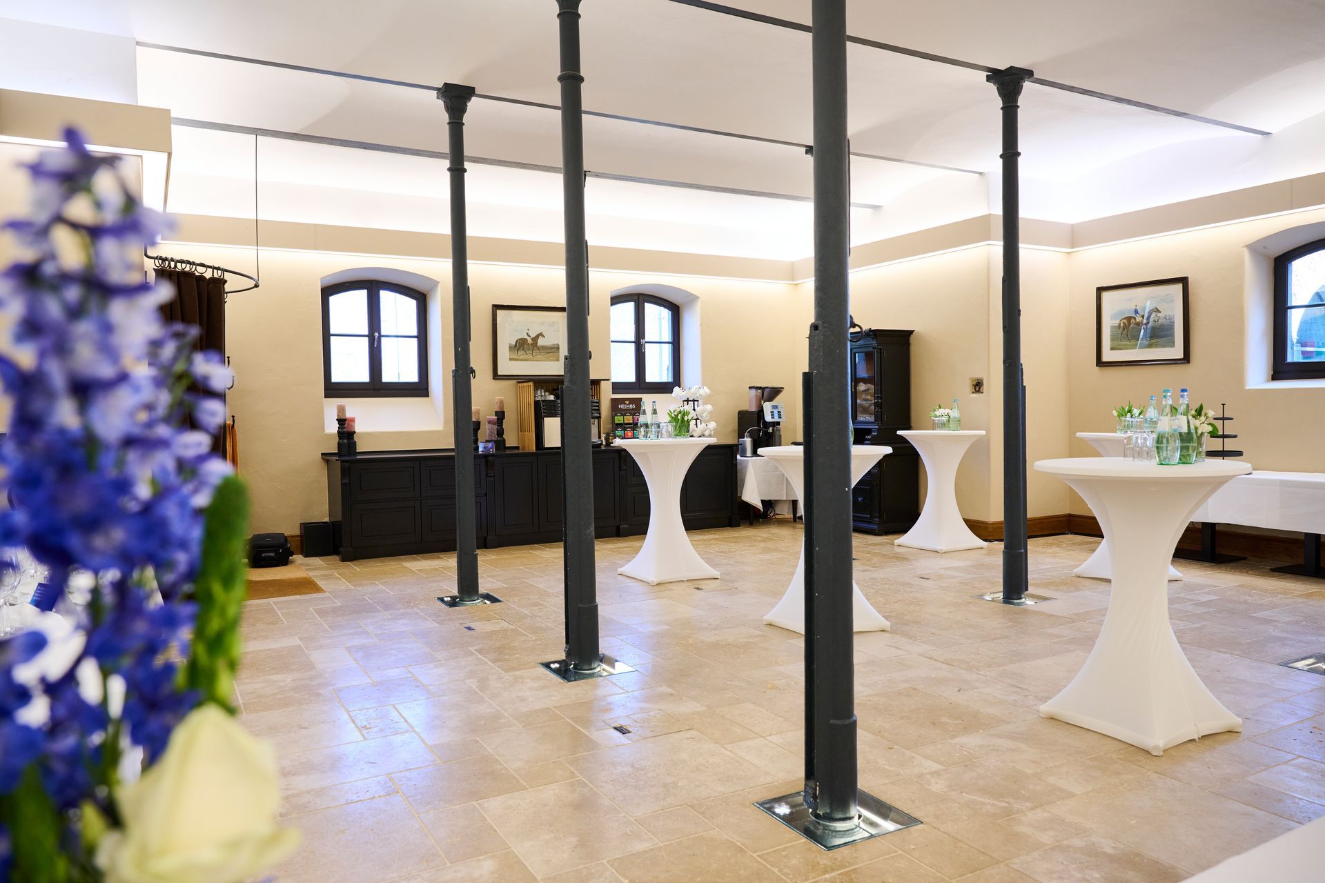 A spacious room with stone floors, dark columns, and white-clothed bar tables set up for an event.