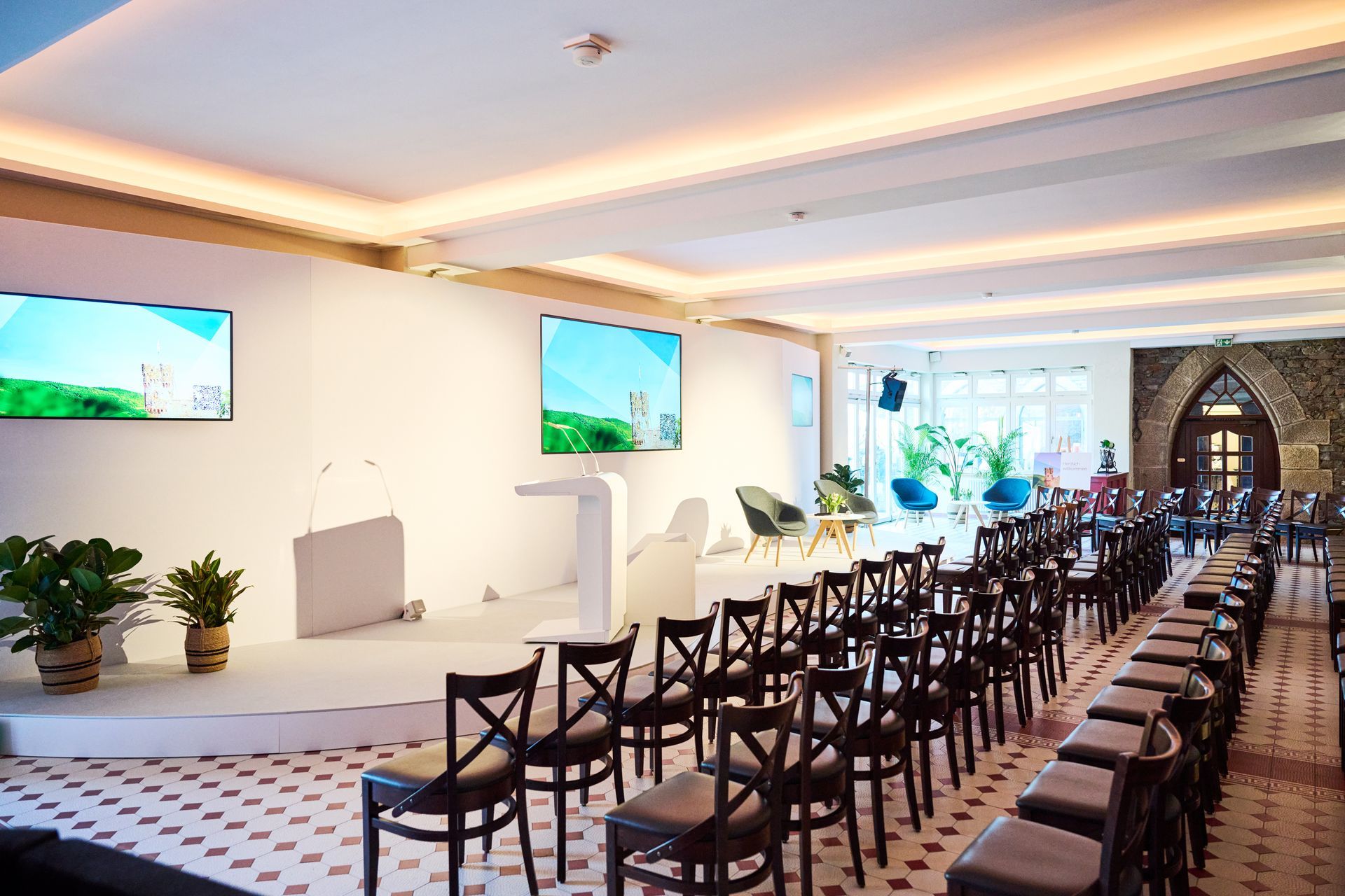 An empty conference room with rows of chairs facing a stage equipped with two screens and a lectern, bathed in warm light.
