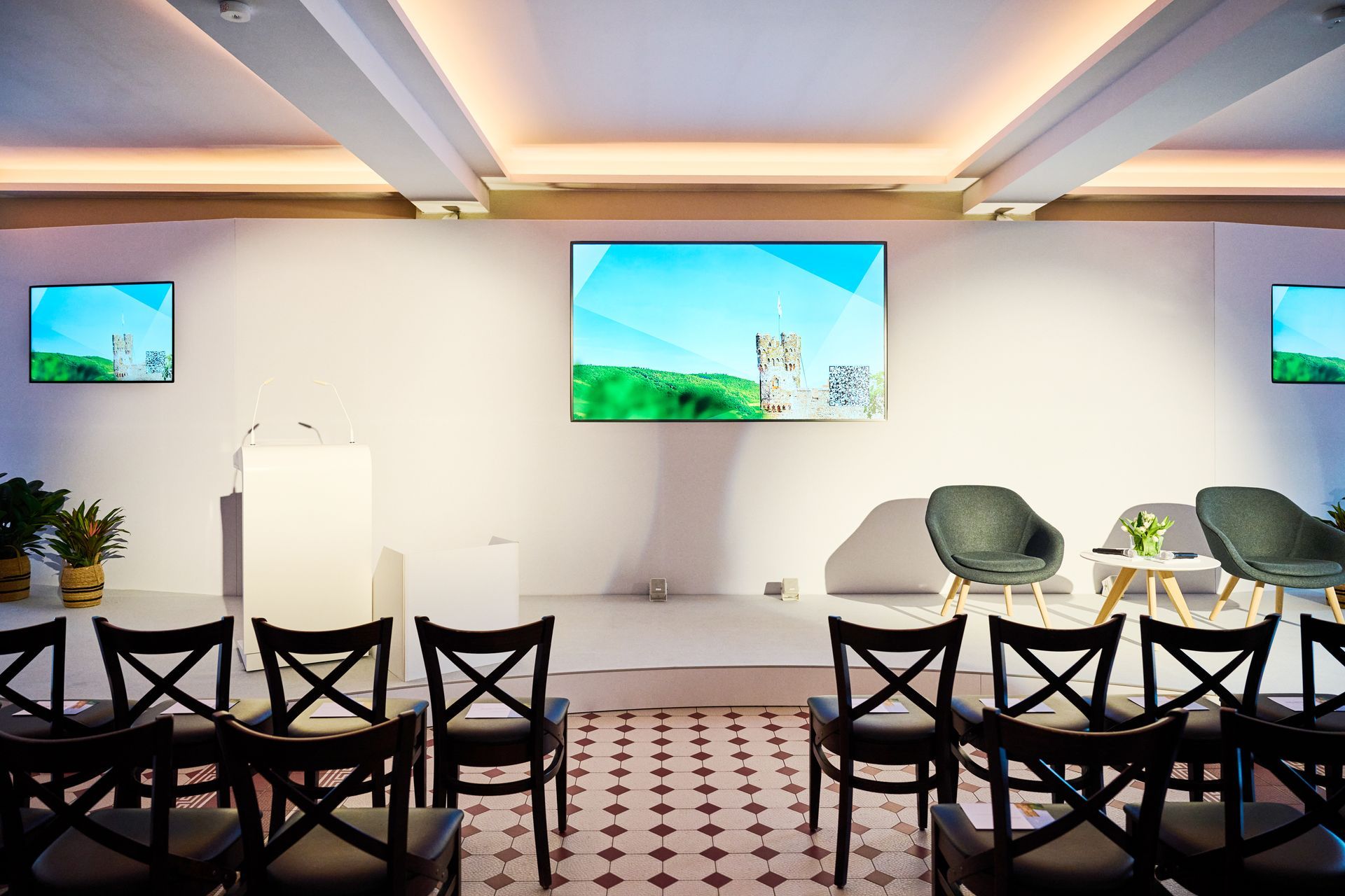 Eine hell erleuchtete Bühne mit Rednerpult, drei Bildschirmen mit Naturaufnahmen und dunklen Stühlen, die für eine Präsentation aufgestellt sind.