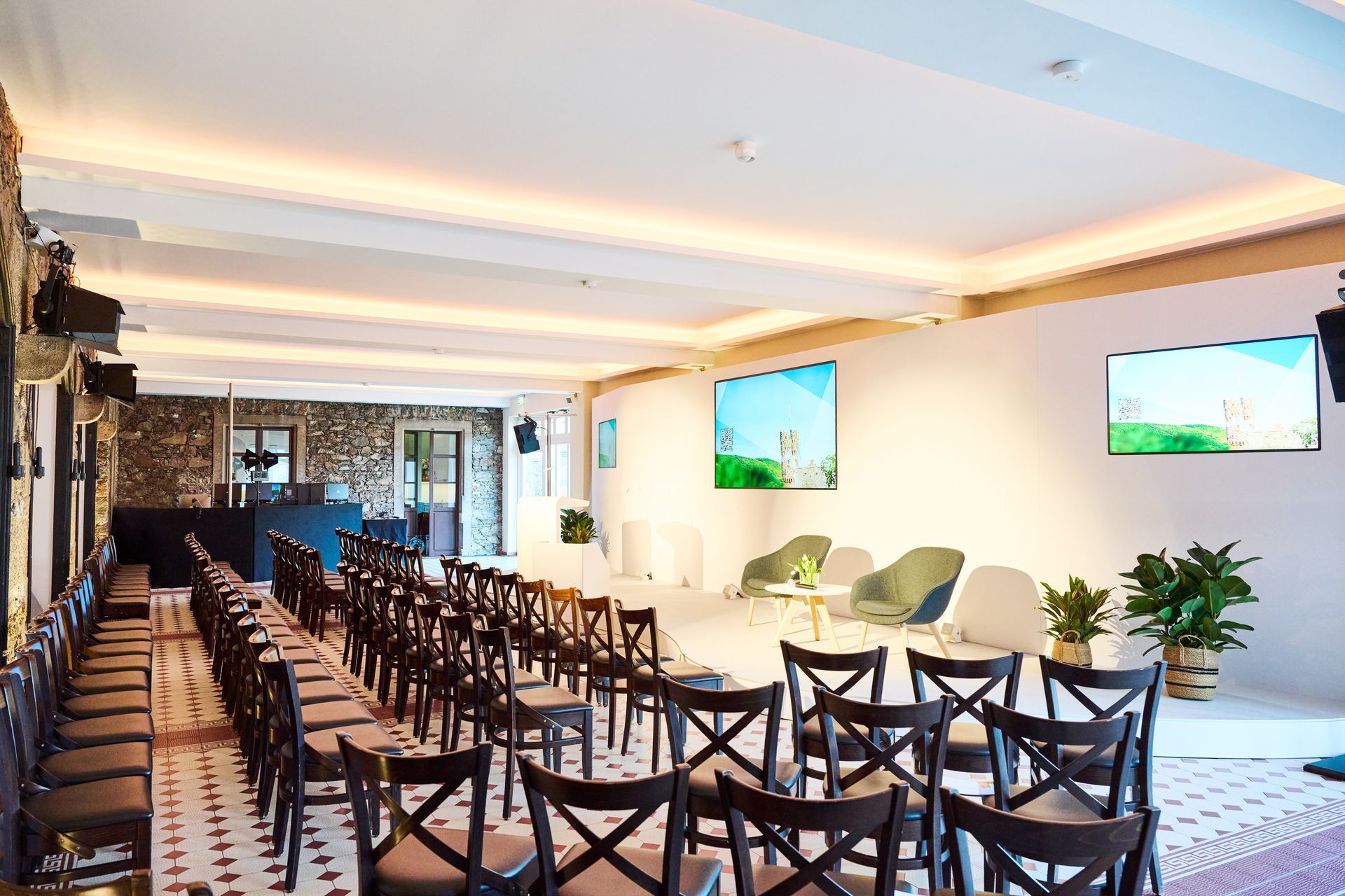 An empty auditorium with rows of dark wooden chairs facing a stage with two green armchairs and two large screens.