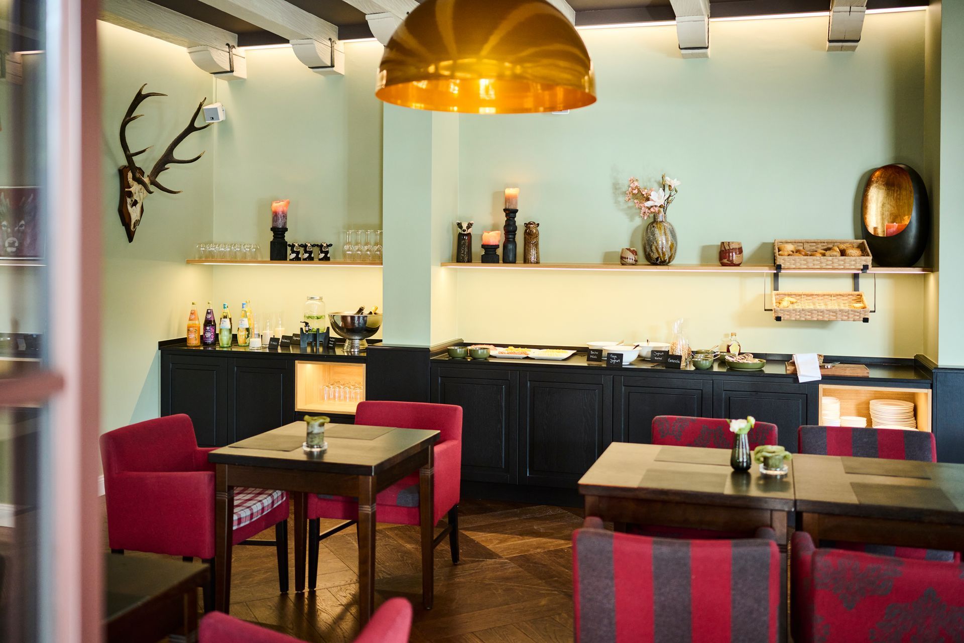 Dining area with tables, red upholstered chairs, and a serving counter stocked with snacks, set against a light green wall.
