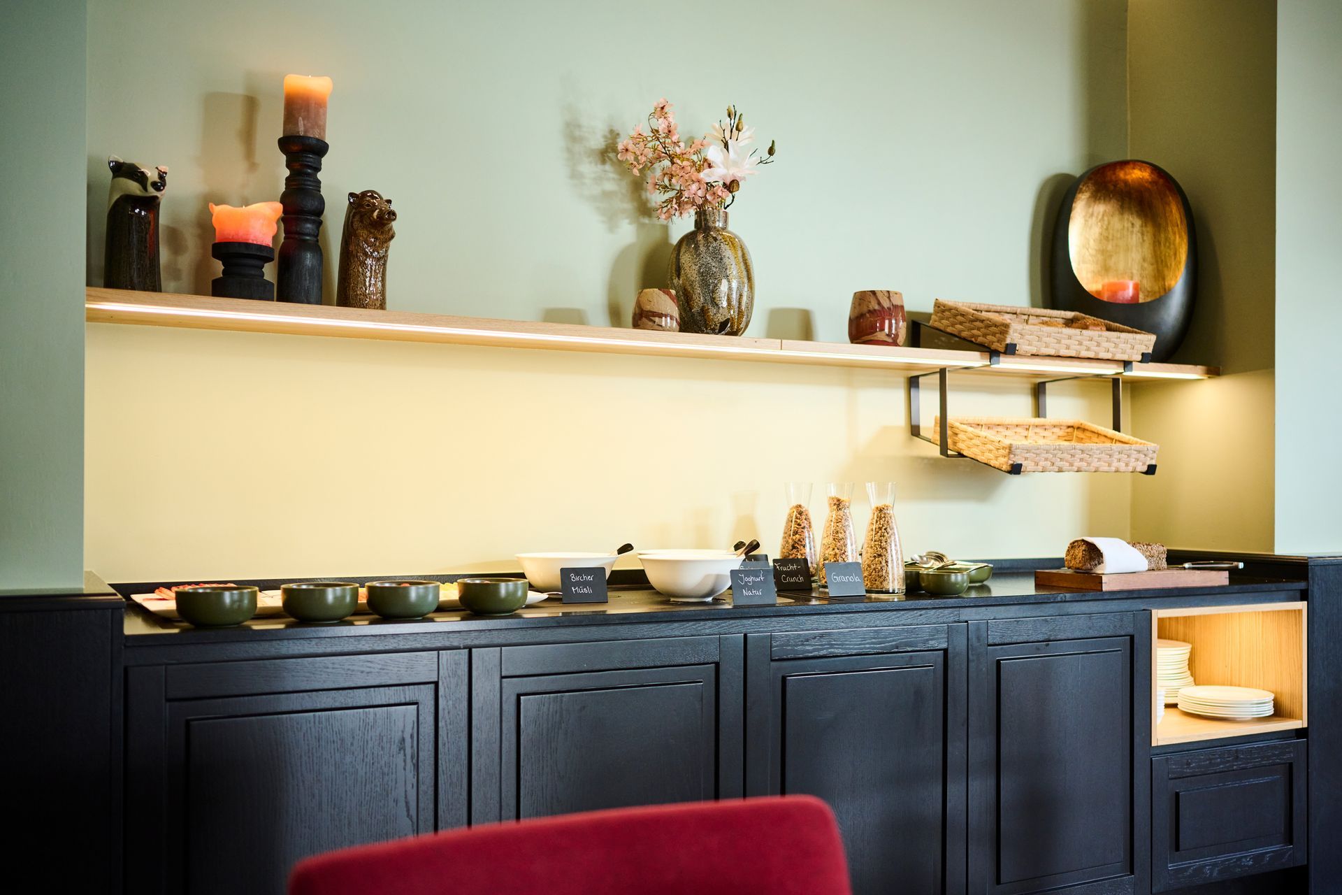 Ein schwarzes Buffet aus Holz mit einem darüberliegenden Regal, auf dem Dekorationen, Kerzen und Küchenutensilien vor einer salbeigrünen Wand präsentiert werden.