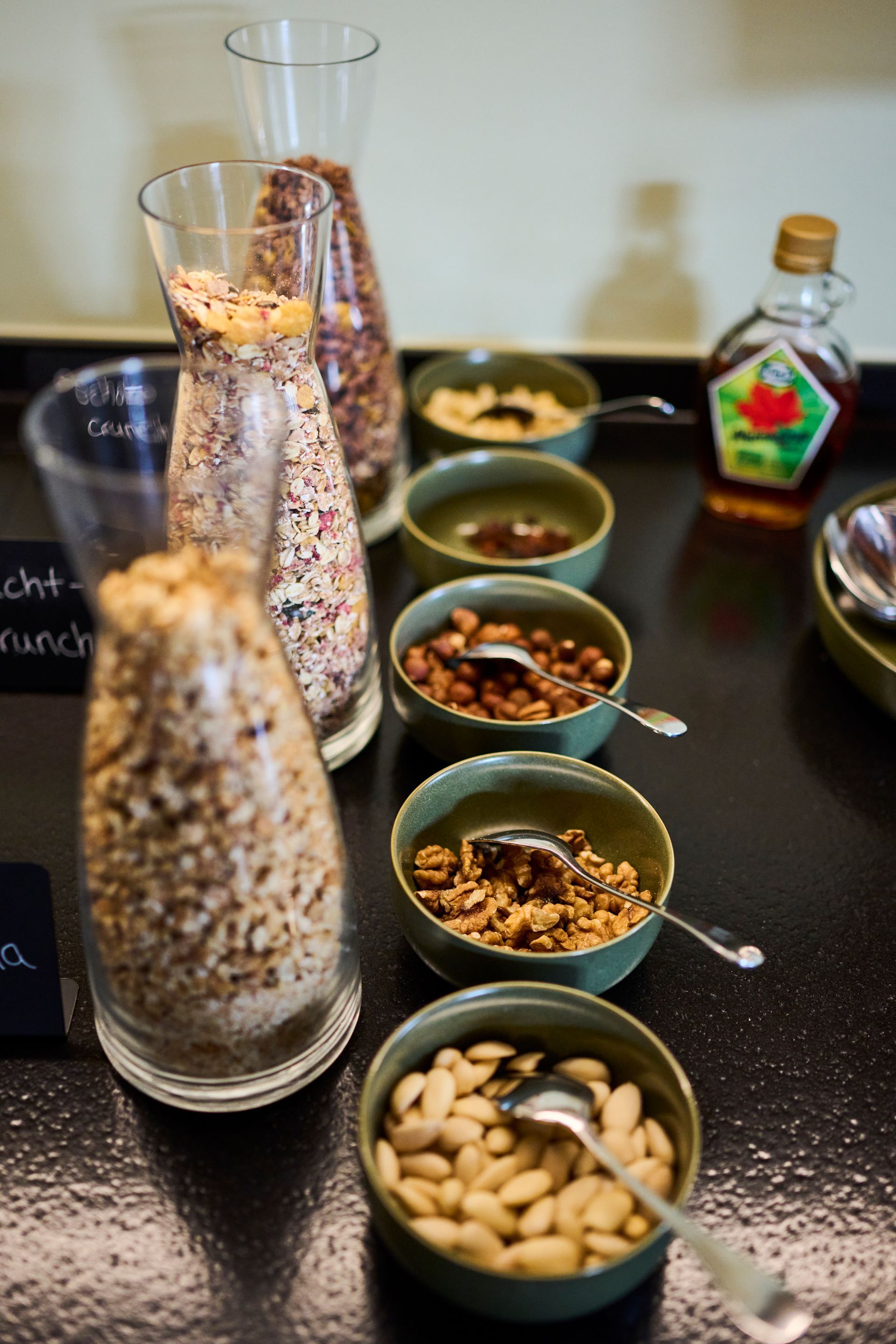 Auf einem schwarzen Tisch war ein Frühstücksbuffet aufgebaut, mit Gläsern voller Müsli, kleinen Schälchen mit Nüssen und Samen und einer Flasche Sirup.