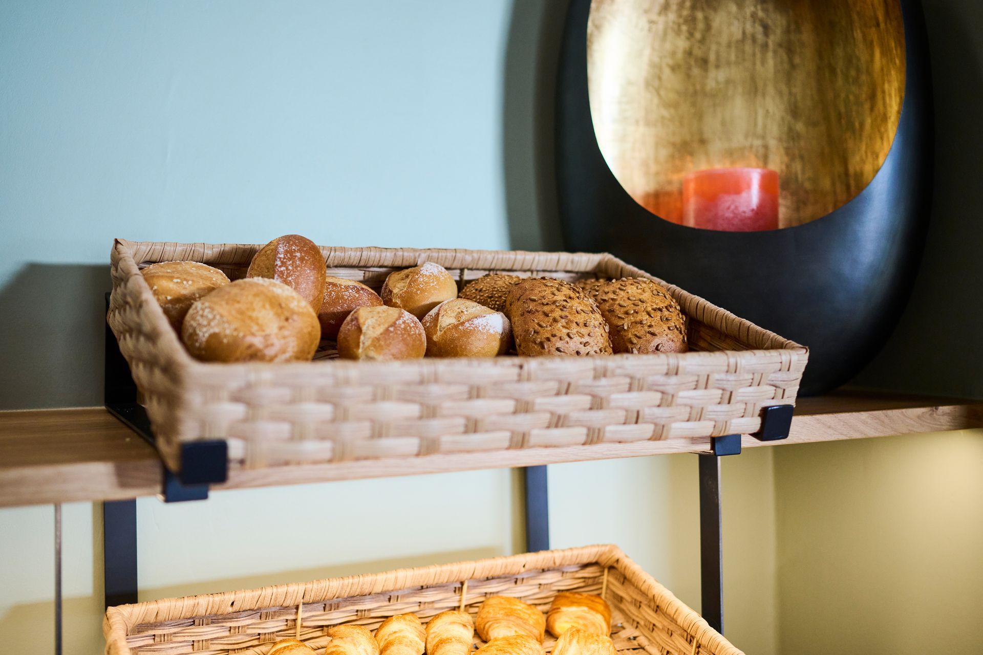 Auf Holzregalen stehen Weidenkörbe gefüllt mit verschiedenen frischen Brötchen, im Hintergrund prangt ein dekorativer goldener Wandakzent.