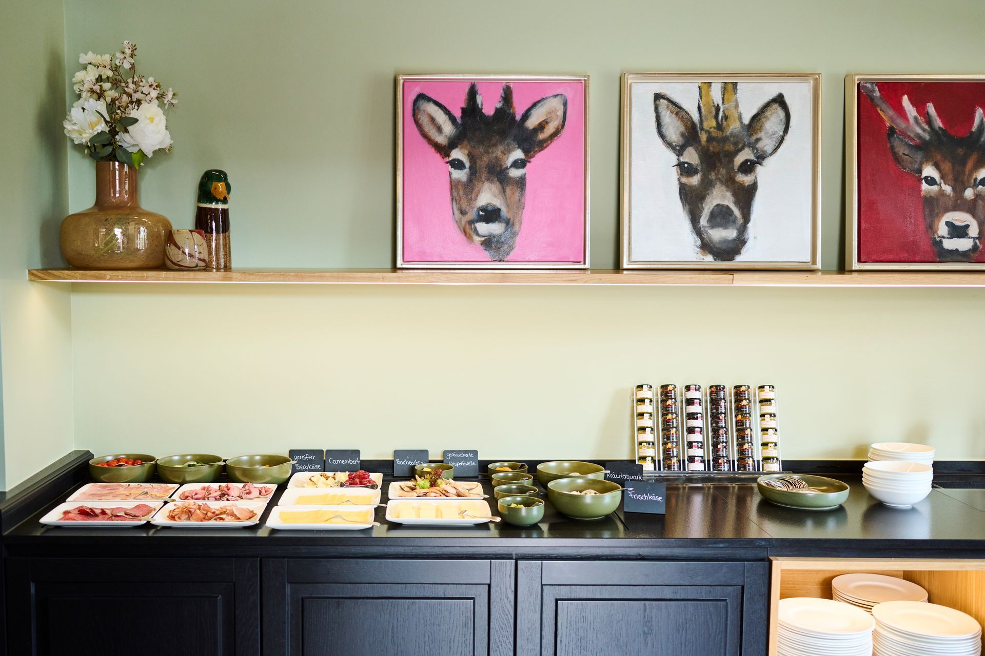 Ein Frühstücksbuffet mit Fleisch, Käse und Schüsseln auf einer schwarzen Theke, unter einem Regal mit Hirschporträts und einer Vase.